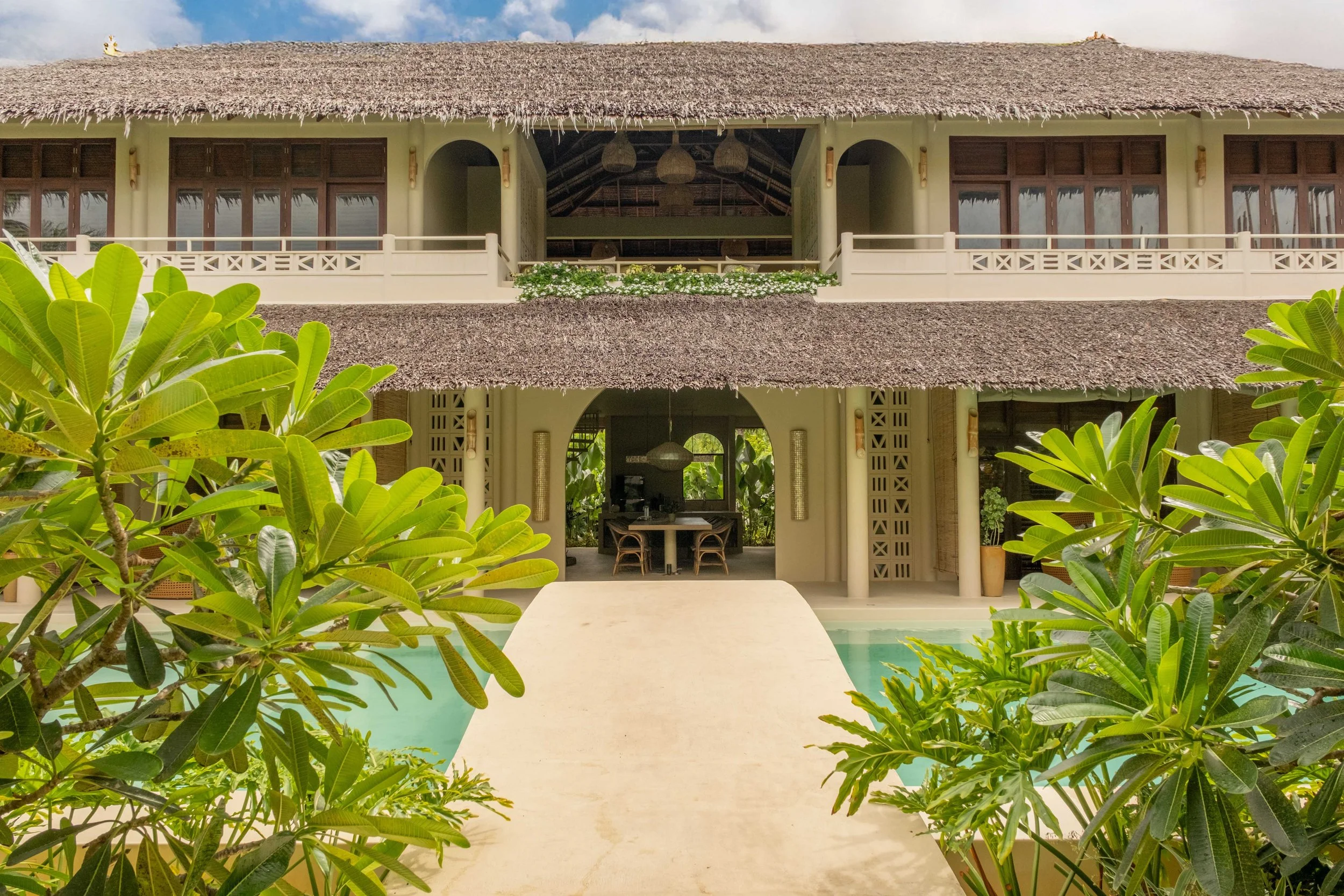 Tropical villa with thatched roof, surrounded by lush green plants, featuring an open balcony and pool area.
