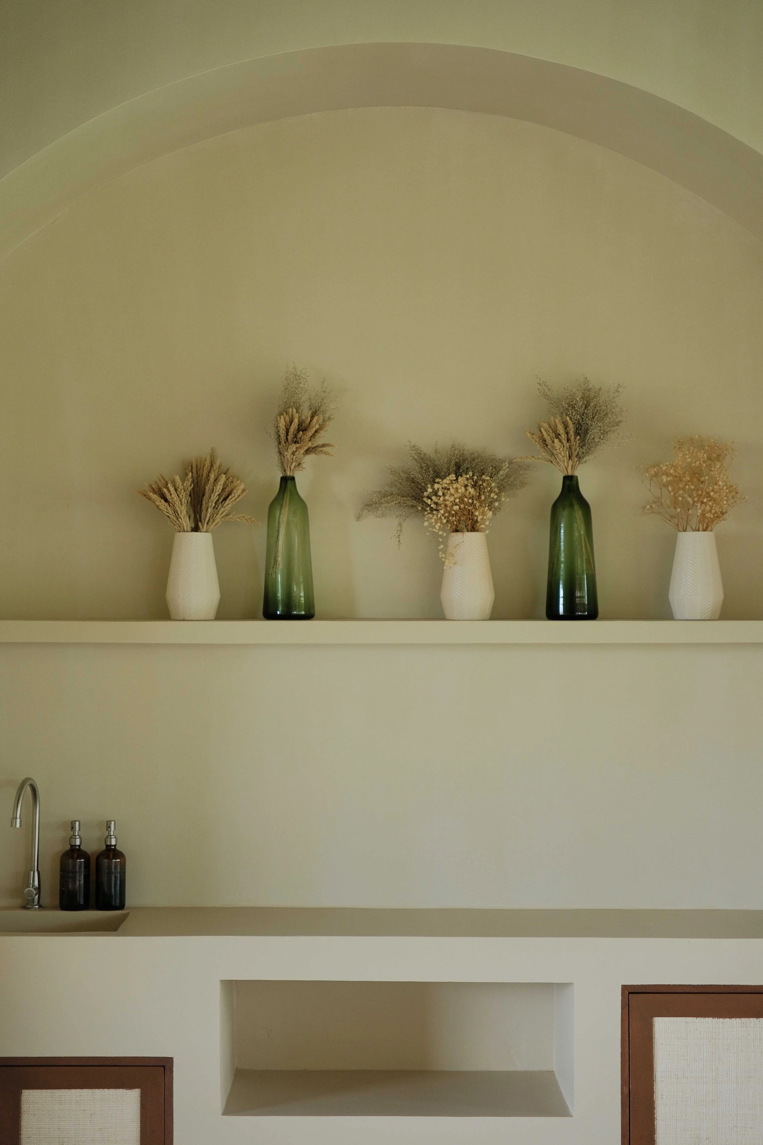 Minimalist kitchen with neutral-toned decor, featuring dried plants in green and white vases on a shelf, a faucet, and brown bottles on the countertop.
