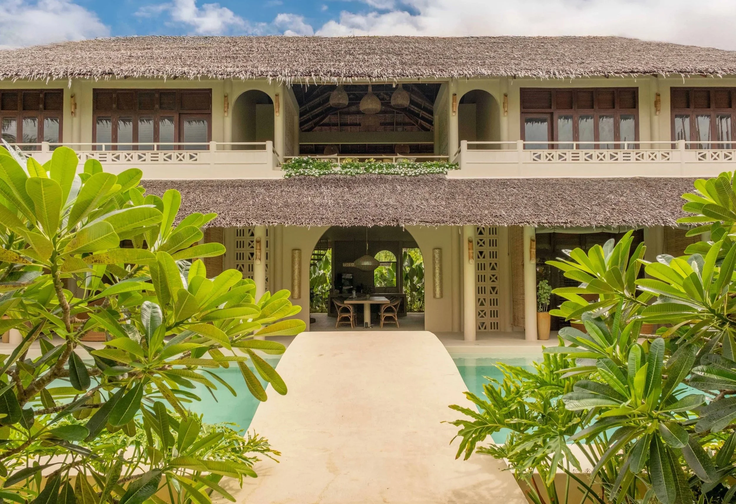 A two-story tropical house with a thatched roof, large windows, and a balcony. Green foliage surrounds a pool area in the foreground.