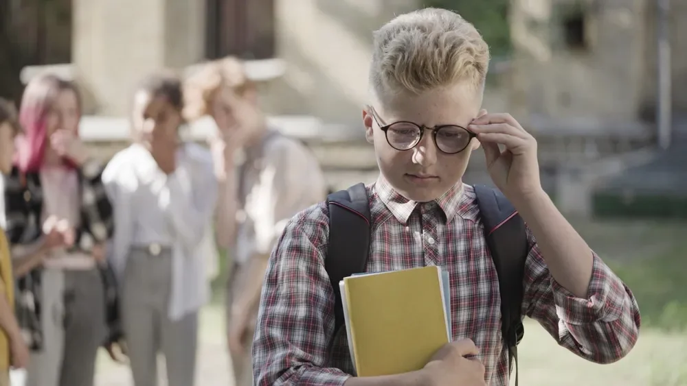 A sad school-aged boy holding books and adjusting his glasses while a group of children in the background appear to whisper and laugh at him.