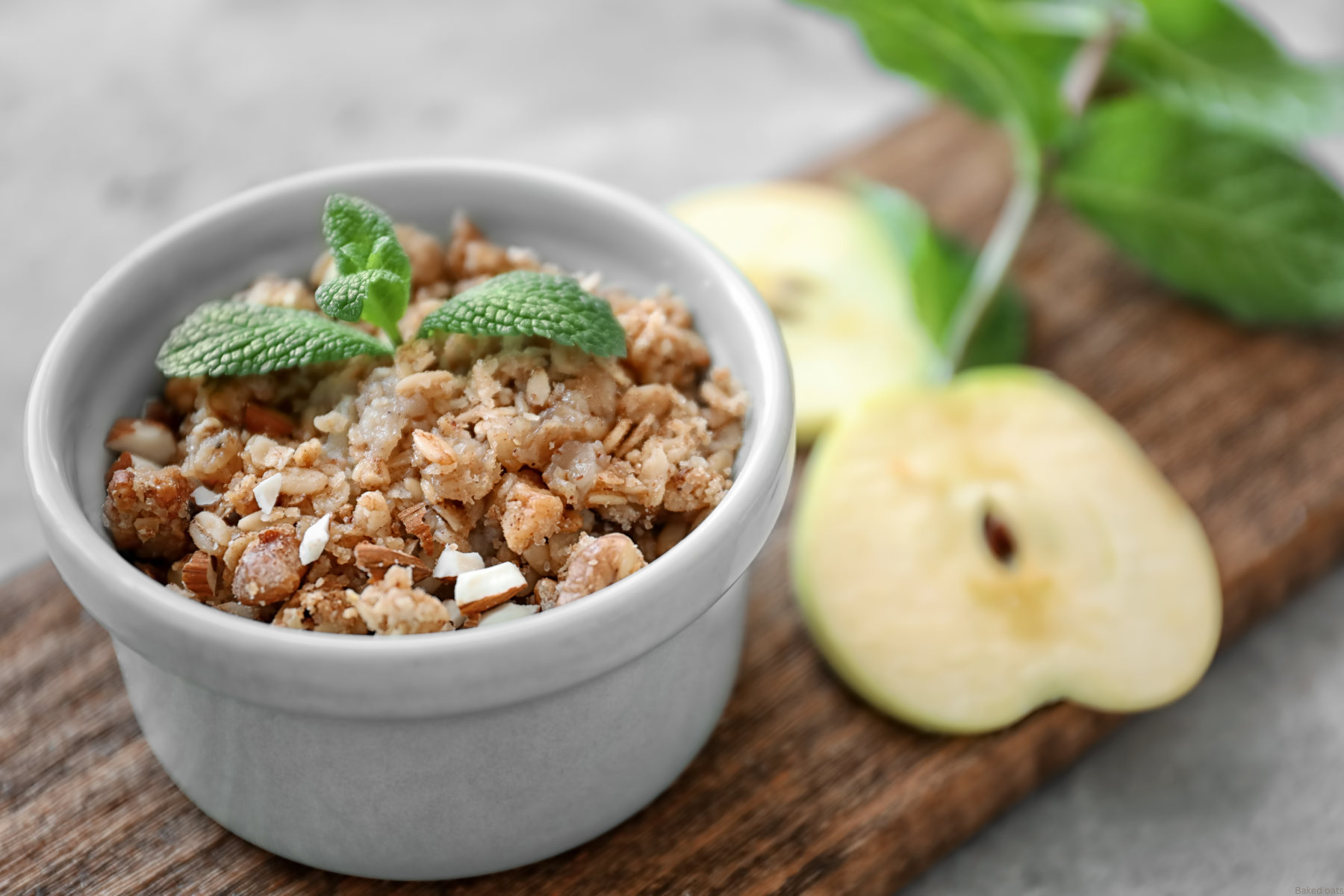 baked oats in a dish with mint leave on top, sliced apple on the side. On a wooden board.