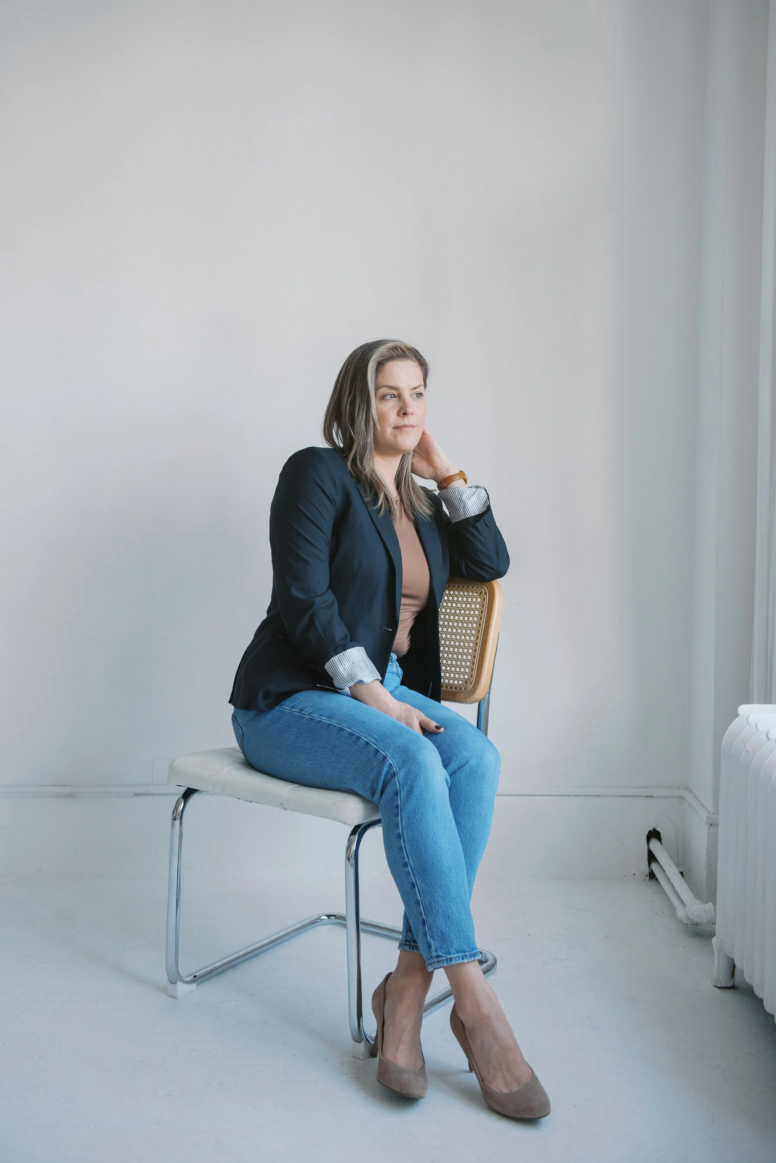 A woman sitting on a white chair with her legs crossed, looking off into the distance, in a room with white walls and a plain floor. She is wearing a black blazer, beige top, blue jeans, and beige high heels. She has shoulder-length brown hair and is touching her neck with her right hand.