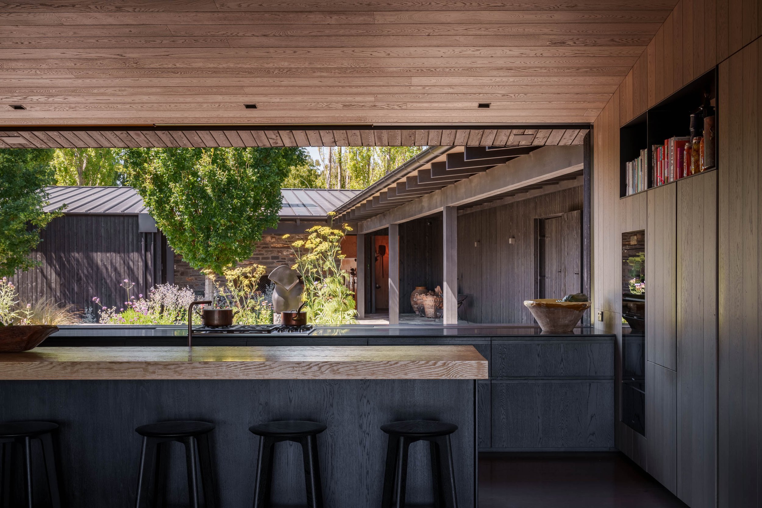 Modern kitchen with wood accents, black cabinetry, a view of a garden outside, and three black stools at the kitchen island.