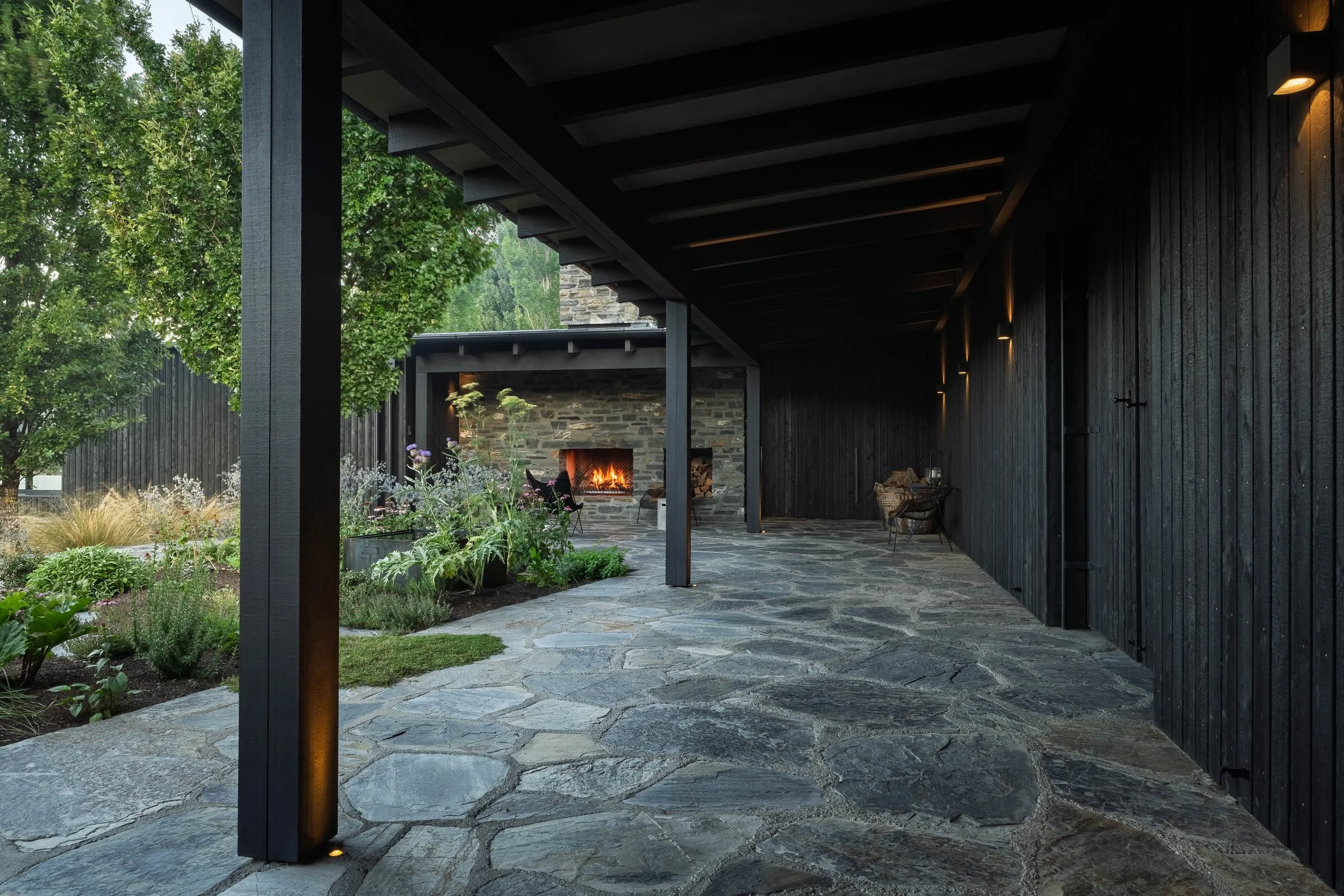 A covered outdoor patio area with a stone floor, black wooden walls, and a fireplace with a fire burning inside. There are outdoor chairs, potted plants, greenery, and trees visible in the background.