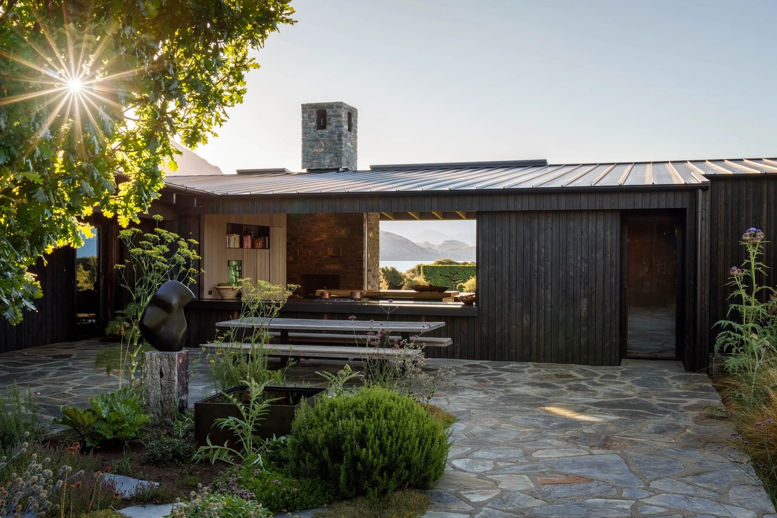 A modern black wooden house with a stone patio, outdoor plants, and a view of mountains and water through an open window, with sunlight shining through a tree.