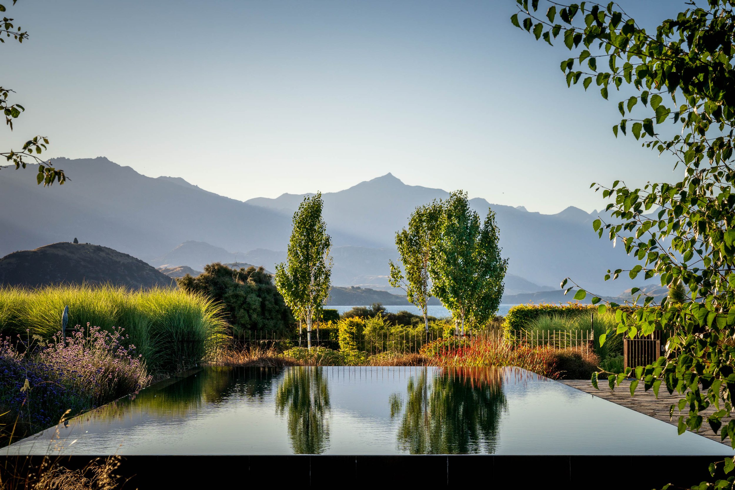 A serene outdoor scene featuring a reflecting pool surrounded by vibrant green plants and trees, with mountainous landscape in the background under a clear sky.