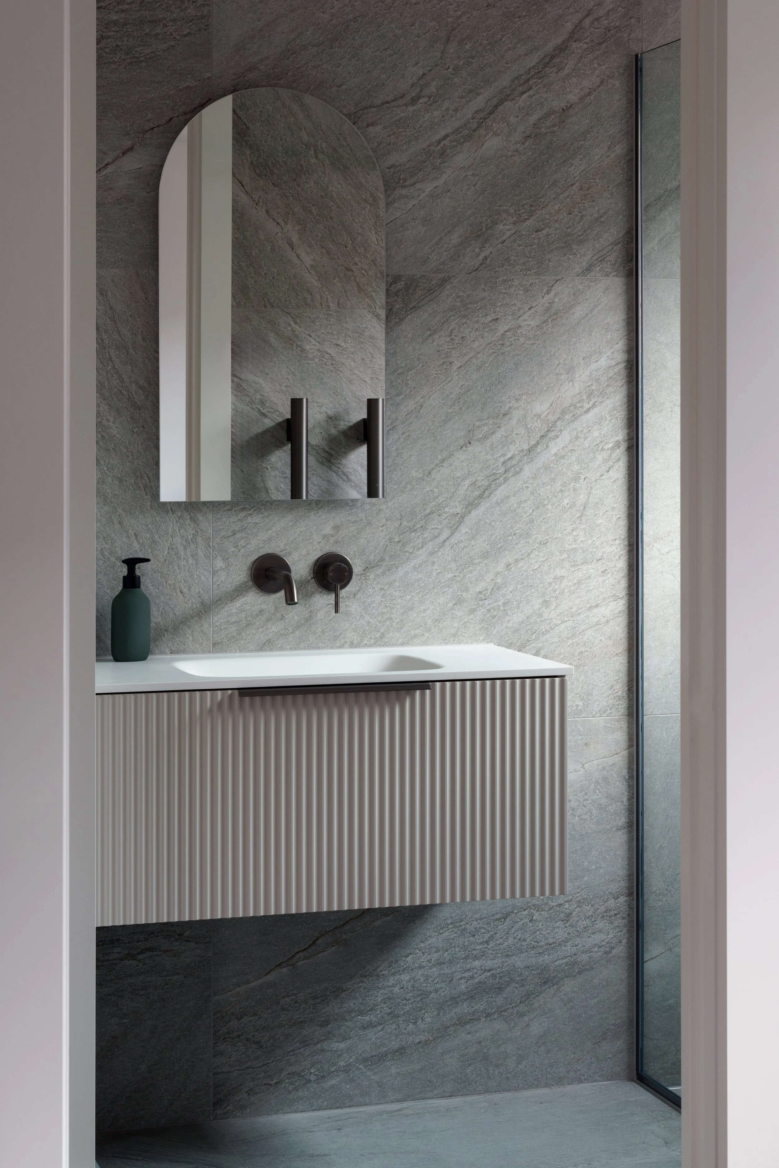 Modern bathroom with a wall-mounted sink, black fixtures, a mirror, a soap dispenser, and a textured gray tile wall.