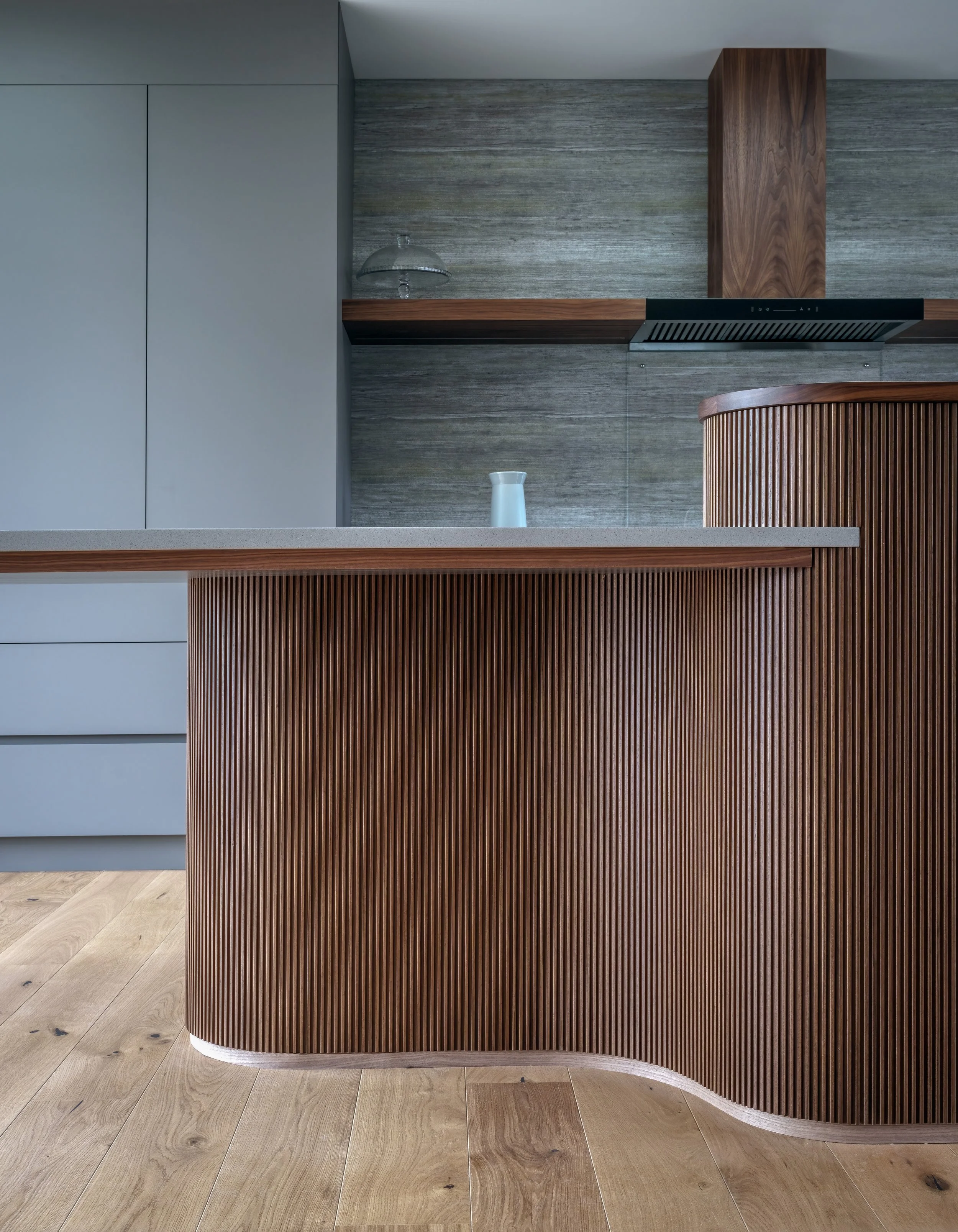 Modern kitchen with a curved wooden island featuring vertical slats, a gray countertop, and a gray and wood-paneled background wall.