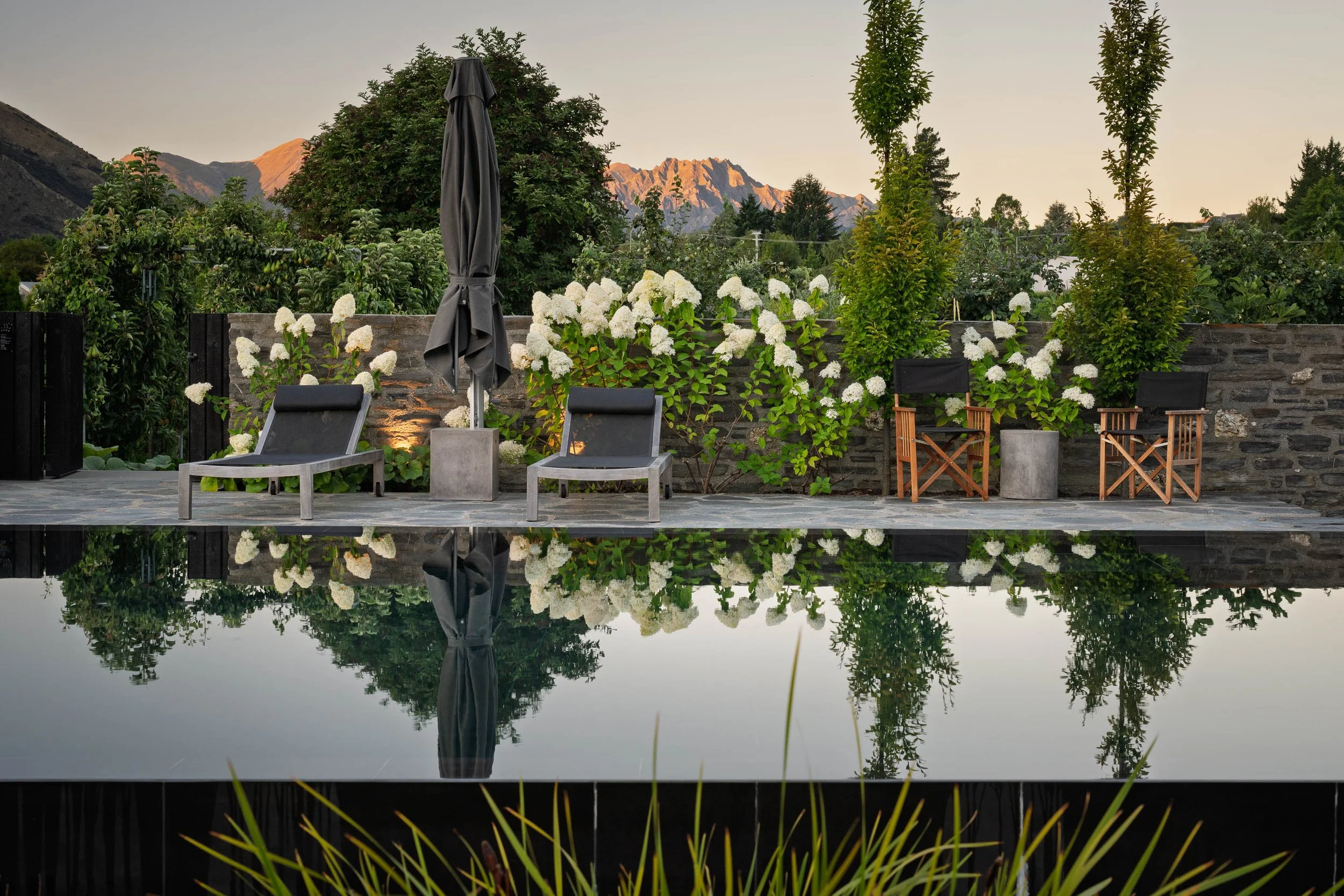 A backyard patio with two lounge chairs and two director's chairs, gazing at a garden with white hydrangeas, tall trees, and mountain range in the background, reflecting in a pool.
