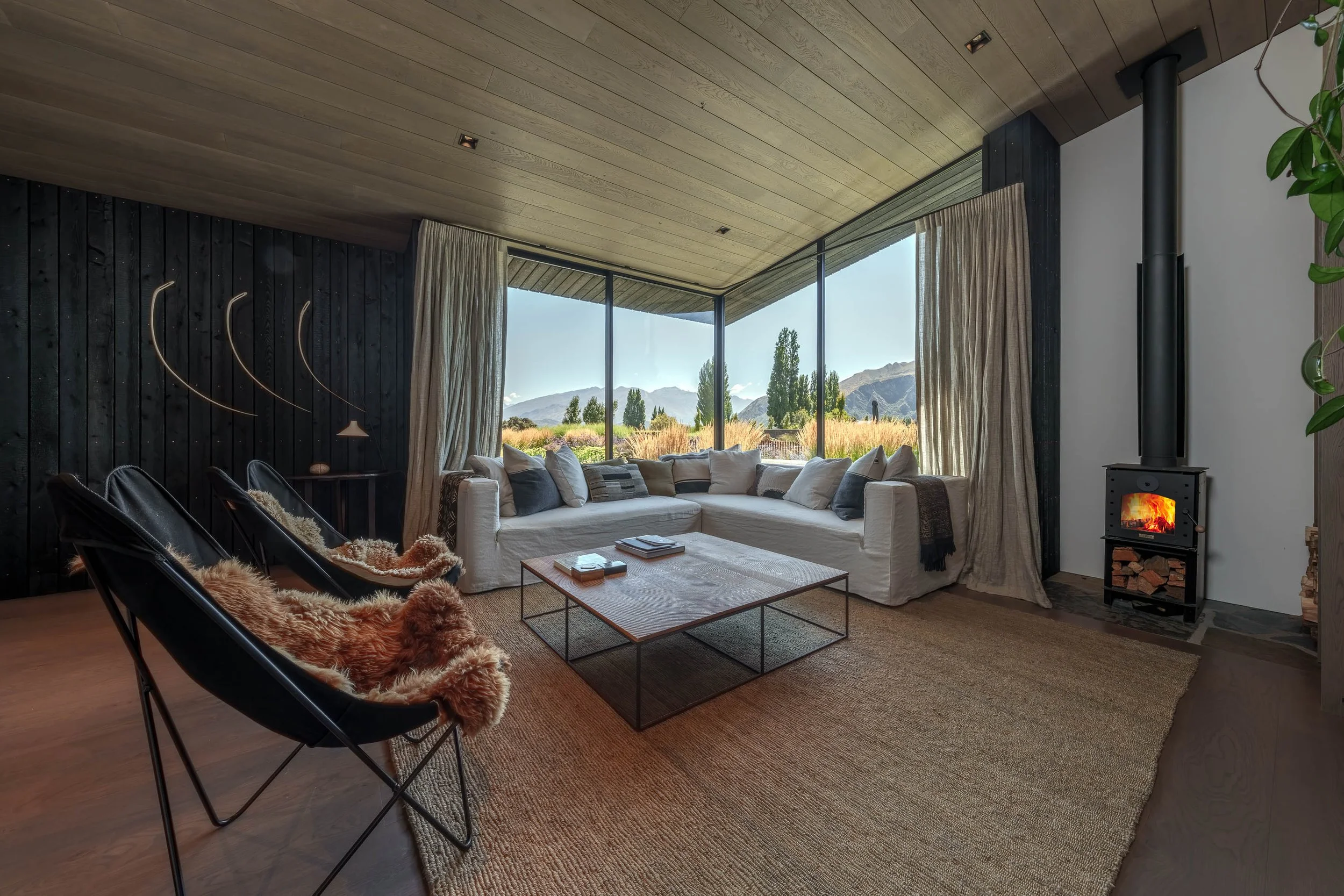 Living room with large window view of mountains and field, white sectional sofa with pillows, wood stove with fire, black chairs with fur throws, coffee table, textured rug, and wooden ceiling.