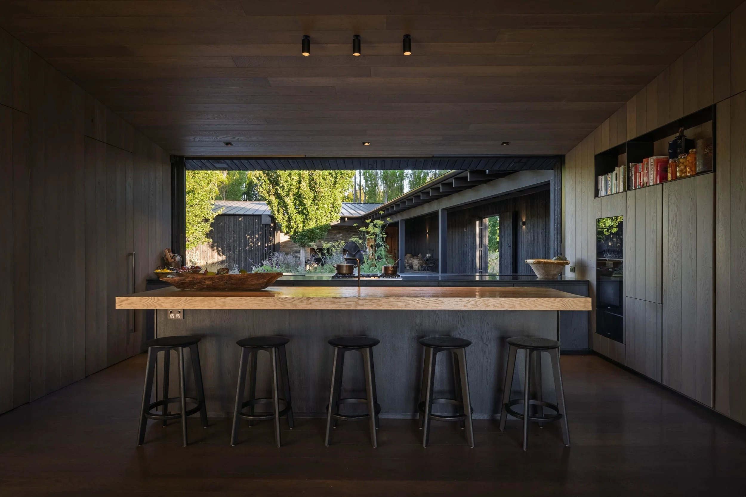 Modern kitchen with dark wood paneling, a large window, a kitchen island with a light wood countertop, five black stools, and built-in cabinets and shelves. Outside garden visible through the window.