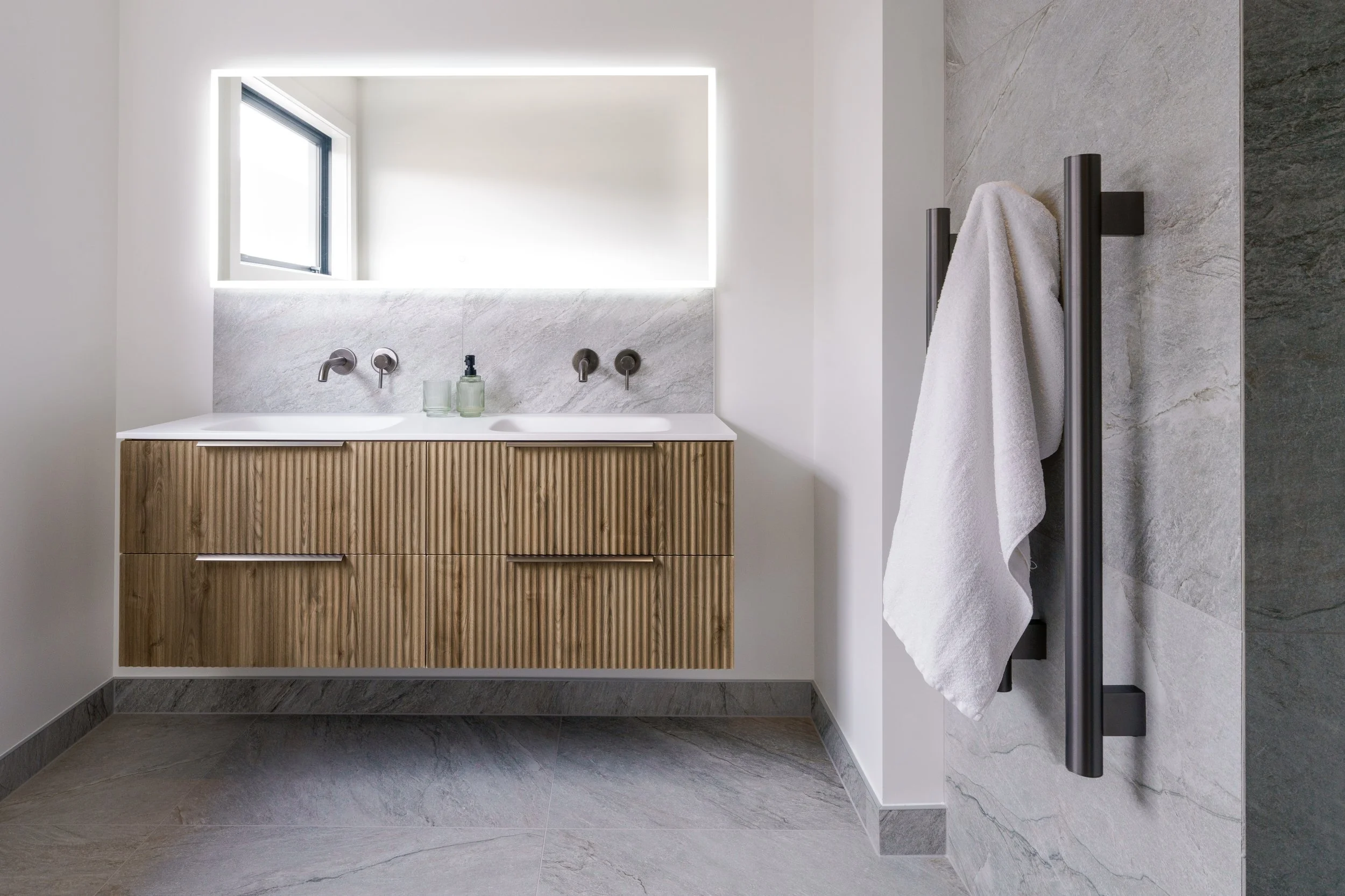 Modern bathroom with a floating wooden vanity, double sinks, and a large illuminated mirror above. A small window beside the mirror lets in natural light, and a white towel hangs on a black vertical towel rack on the right. The bathroom features gray tiles on the floor and wall.
