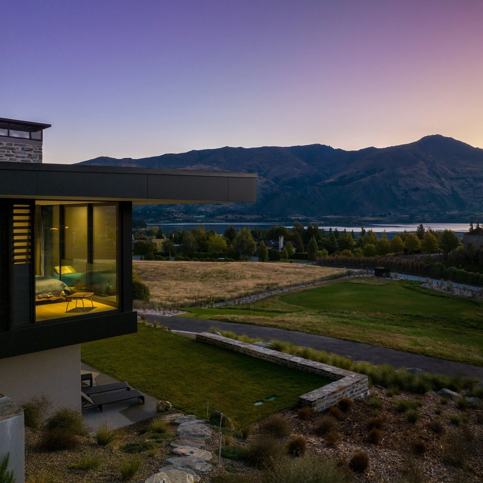 Modern house with a large glass window showing a bedroom inside, overlooking a scenic landscape with mountains, trees, and a grassy field during sunset.