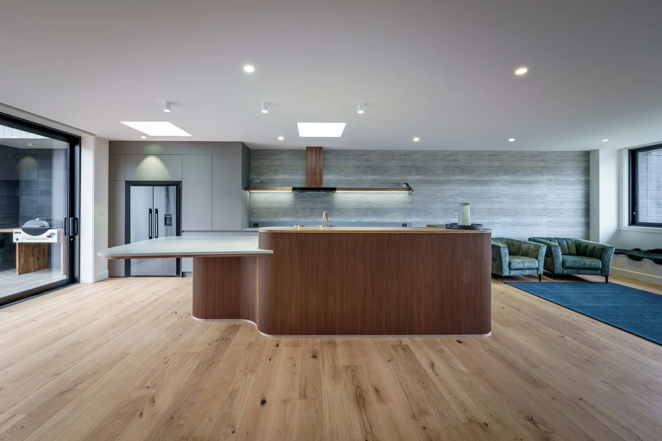 Modern kitchen with wooden island, stainless steel appliances, light wood flooring, gray textured wall, green armchairs, blue rug, and large windows.