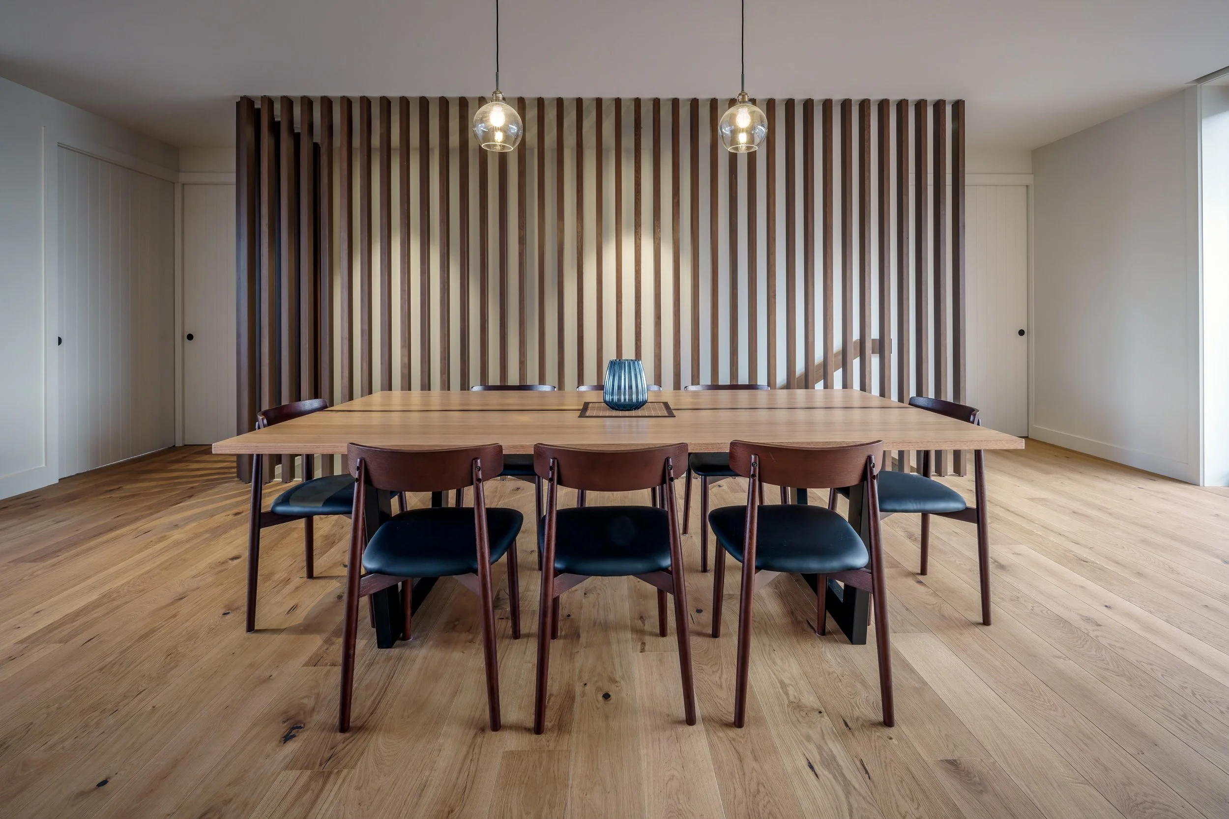 Wooden conference table with eight black and brown chairs in a modern dining room with wooden flooring, a wooden slat wall, hanging pendant lights, and a blue glass vase in the center.