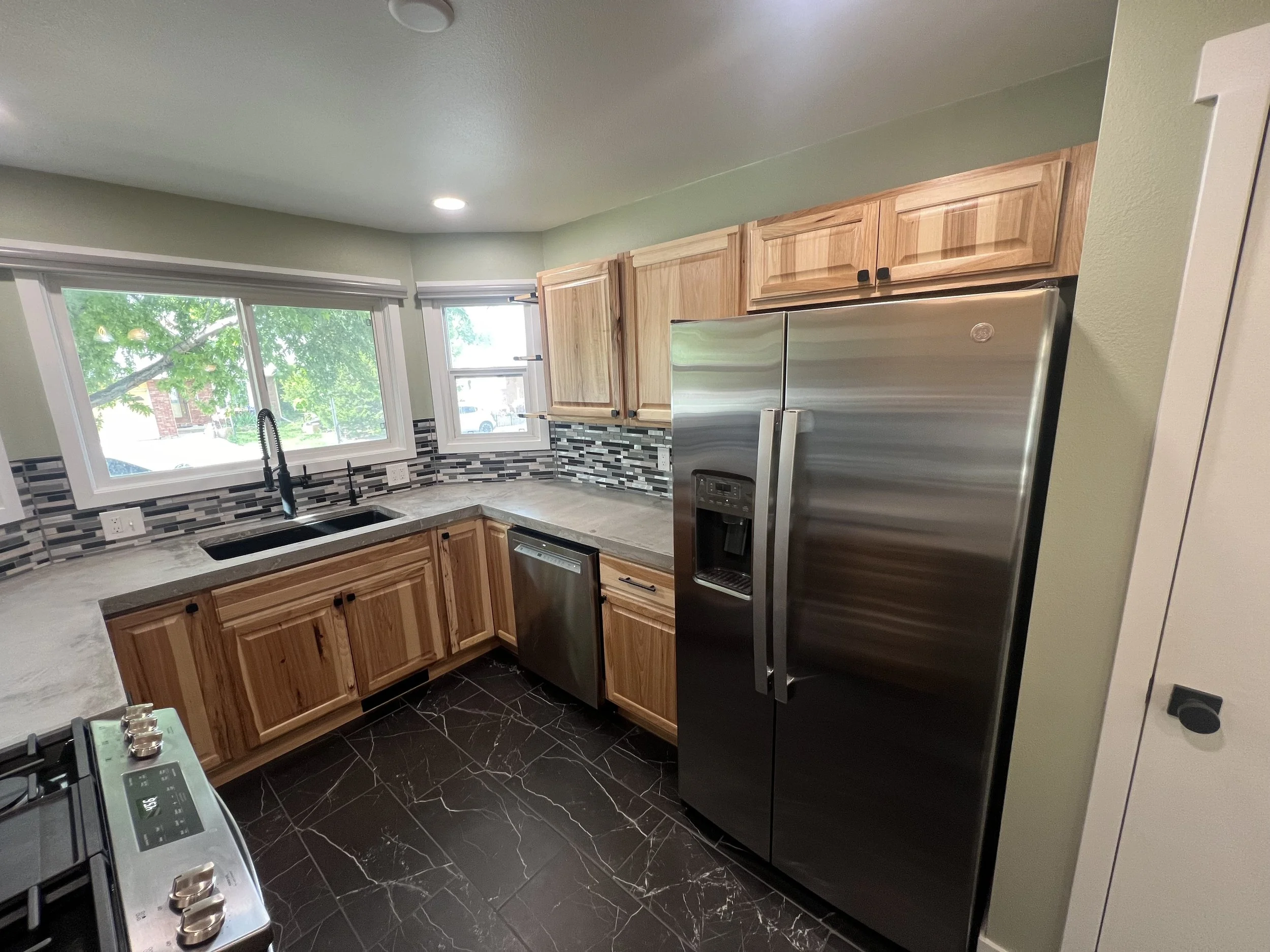 Modern kitchen remodel with wooden cabinets, stainless steel refrigerator, black marble floor, marble countertops, and a window over the sink.