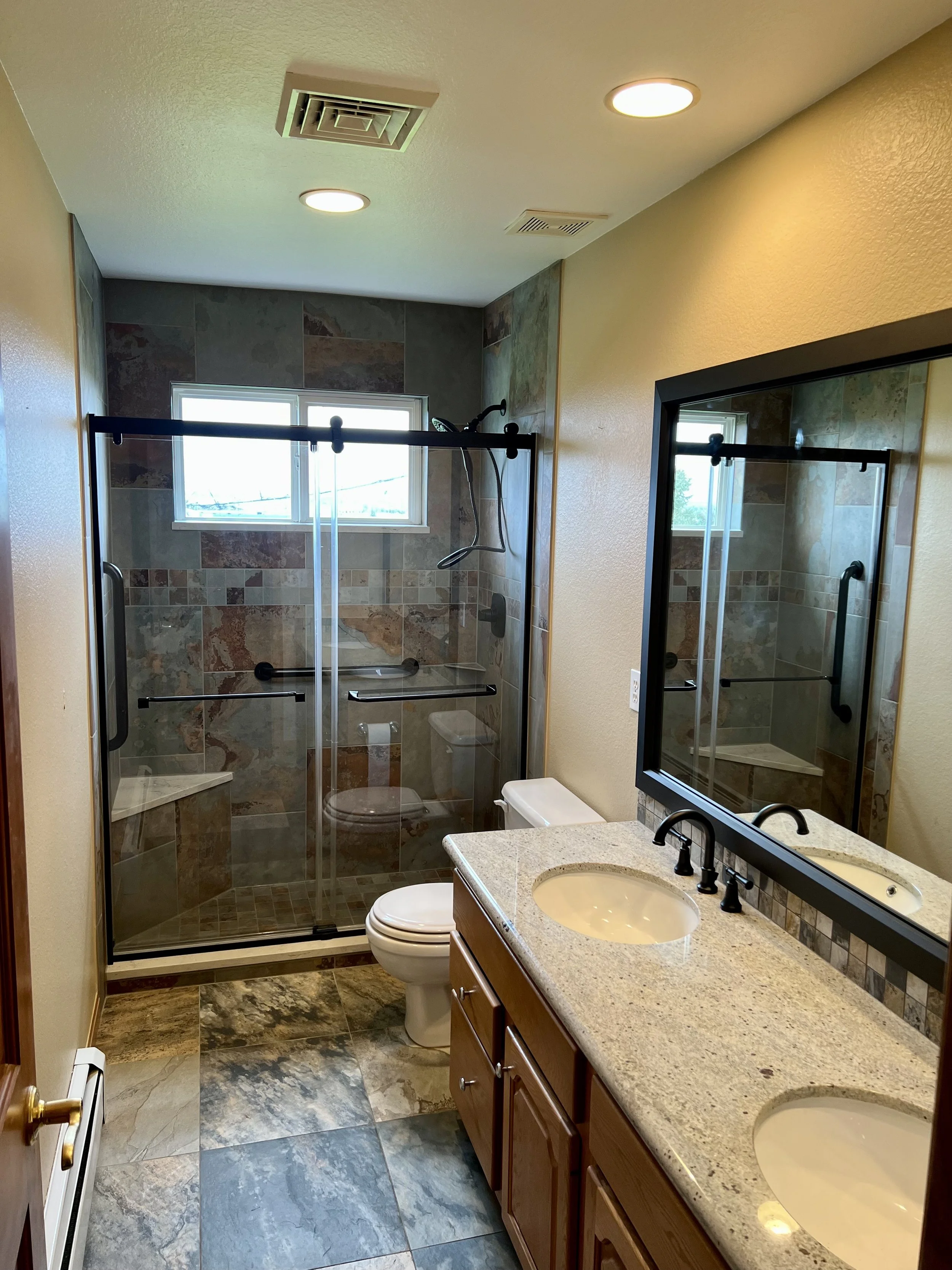 Bathroom remodel with double granite sinks, a wooden vanity, a mirror, a toilet, and a glass-enclosed shower with tiled walls and a window.