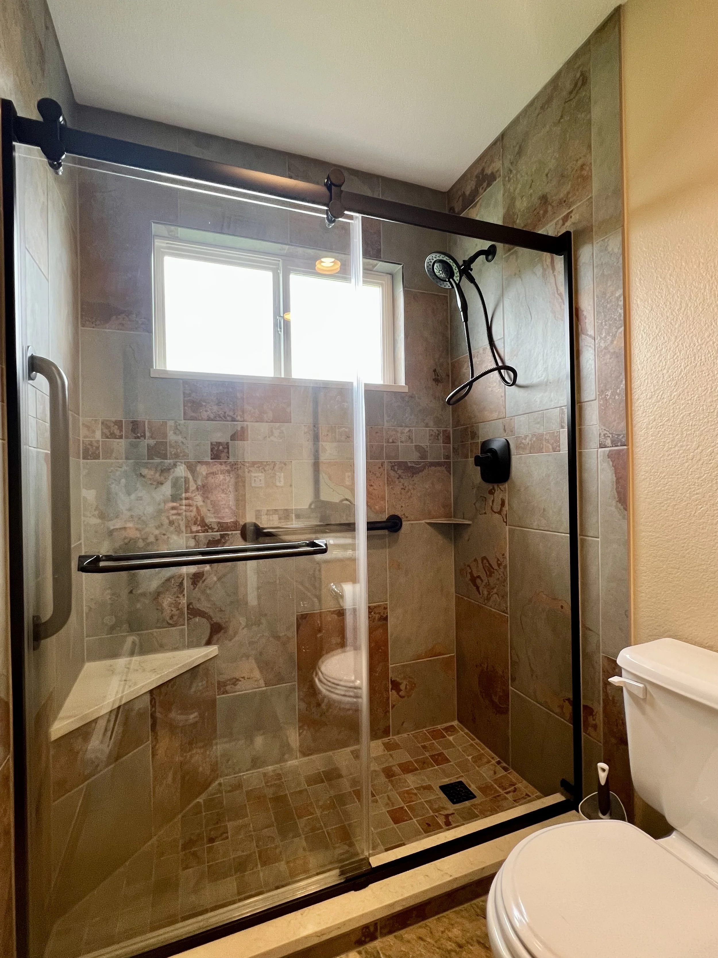 Bathroom remodel shower with clear glass door, tile walls, a small window, and a handheld showerhead inside.