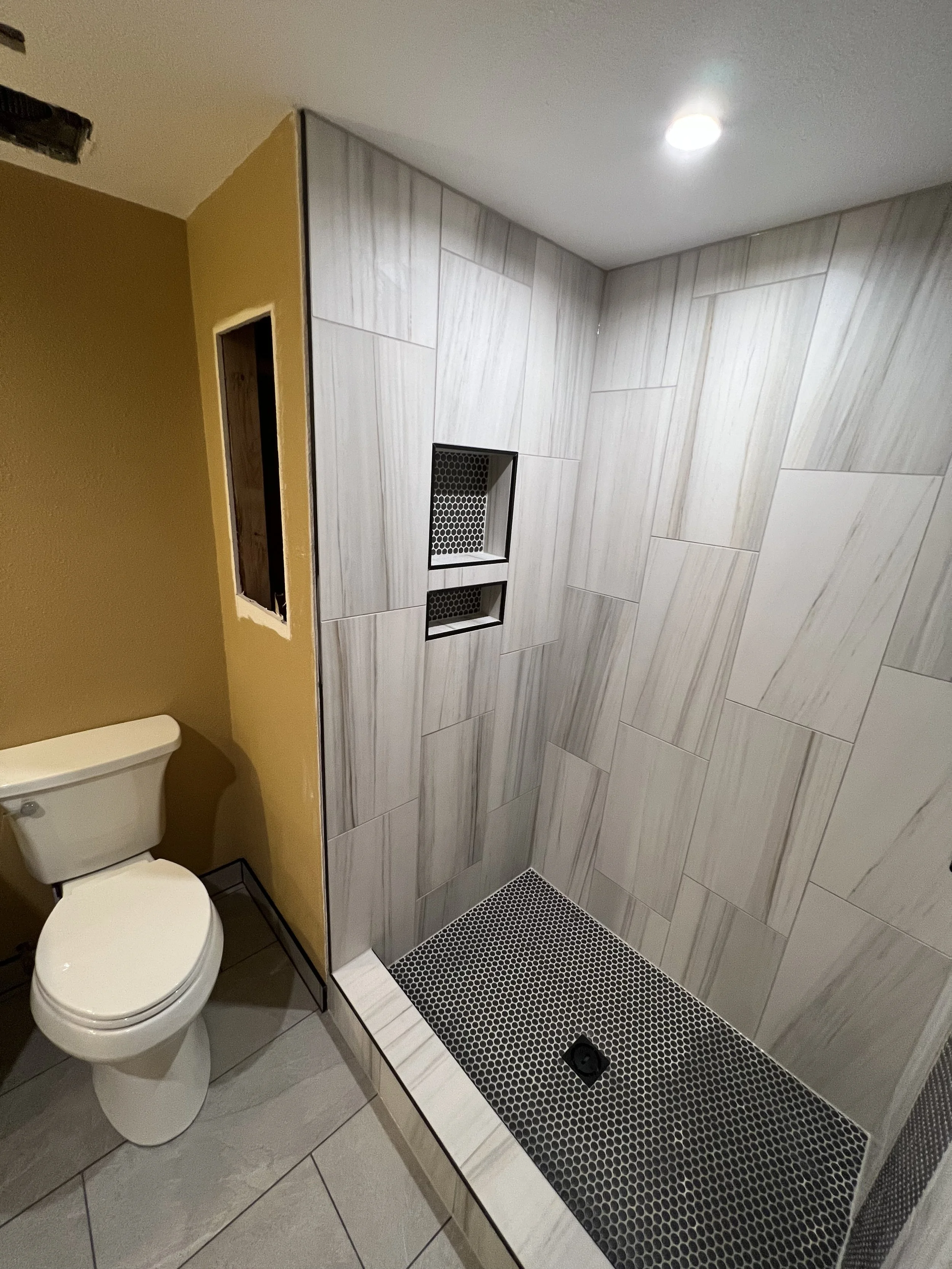 Bathroom remodel with a toilet and a tiled shower stall with two built-in shelves and a drain, featuring beige and white tiles and a ceiling light.