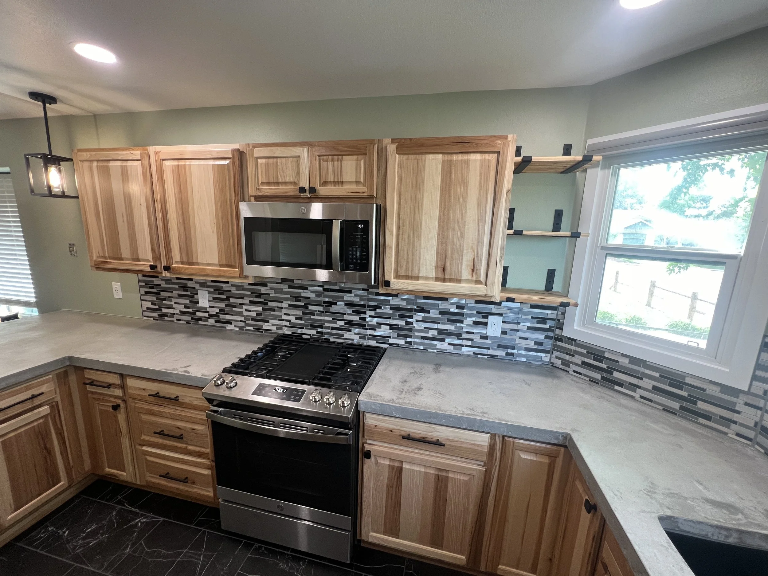 Modern kitchen remodel with wooden cabinets, gray stone countertops, a stainless steel microwave and stove, a mosaic tile backsplash, and a window with a view outside.
