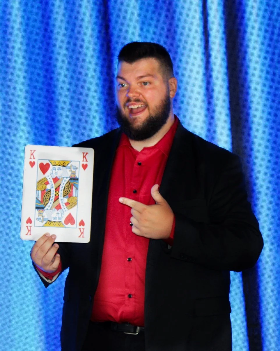 Corporate Magician Seth Dreams with a beard and short hair, wearing a black blazer and red shirt, smiling and pointing at a large King of Hearts playing card he is holding.