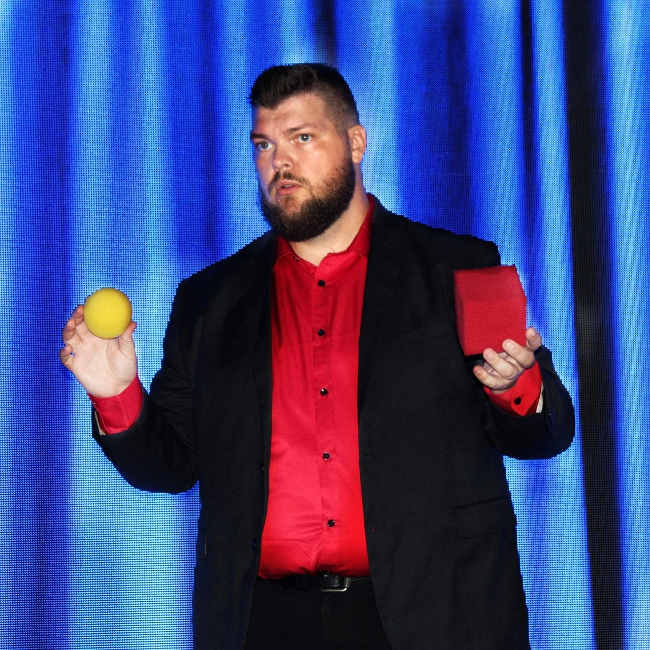 Corporate Magician Seth Dreams with a beard and short dark hair dressed in a black blazer and red shirt holding a yellow ball in his right hand and a red box in his left hand, standing in front of a blue curtain.