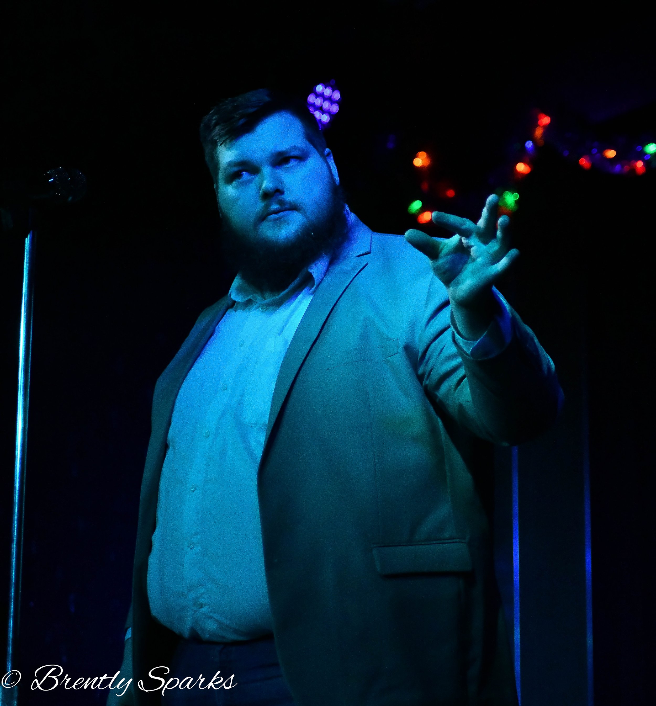 Corporate magician Seth Dreams with a beard in a suit poses with his right hand raised in front of colorful, blurred lights against a black background.