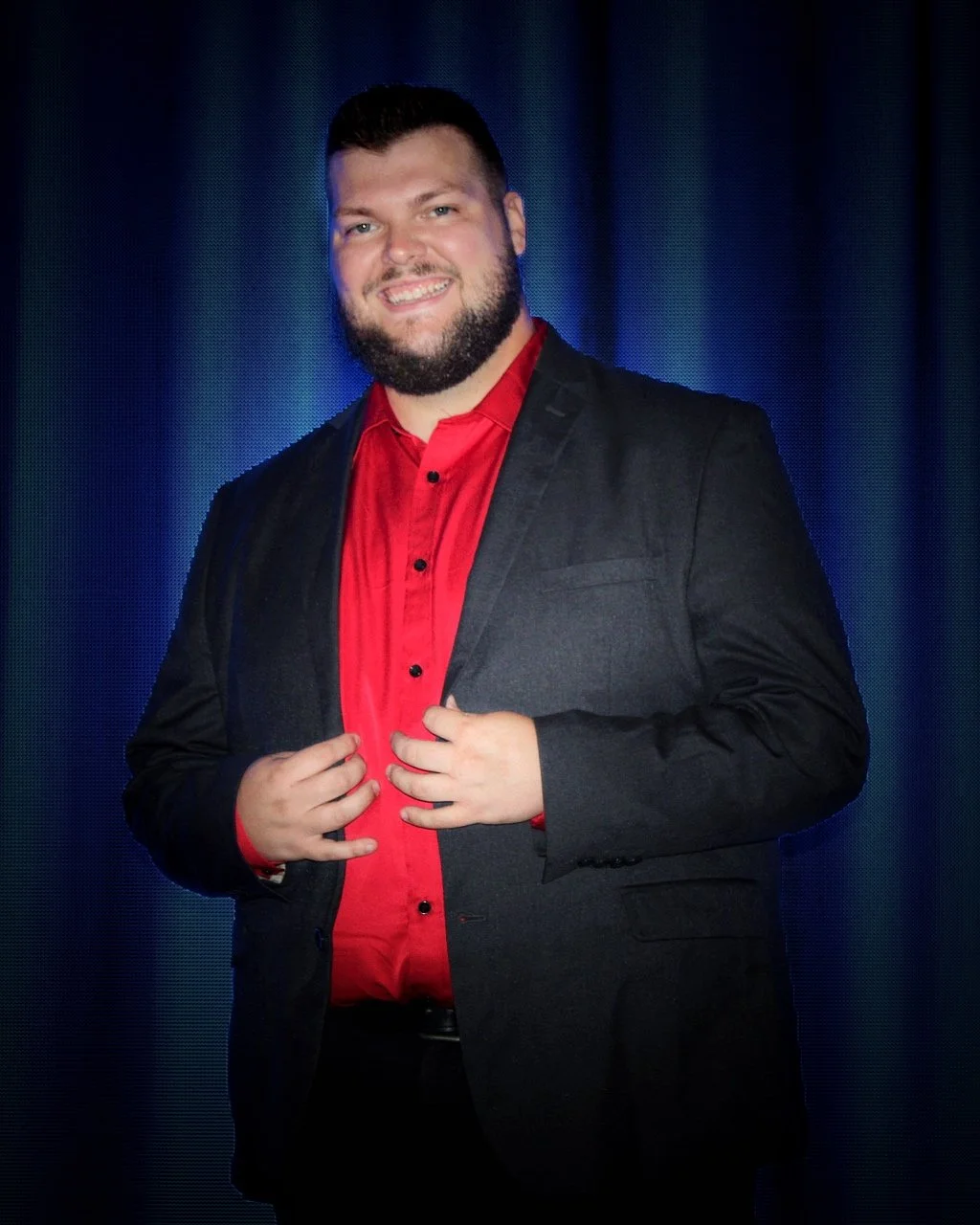 Corporate Magician Seth Dreams with a beard wearing a black suit and red shirt, standing in front of a dark curtain background.