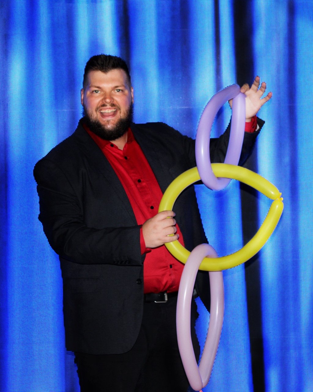 Corporate Magician Seth Dreams with a beard smiling at the camera, holding colorful twisted balloon shapes, standing in front of a blue curtain.