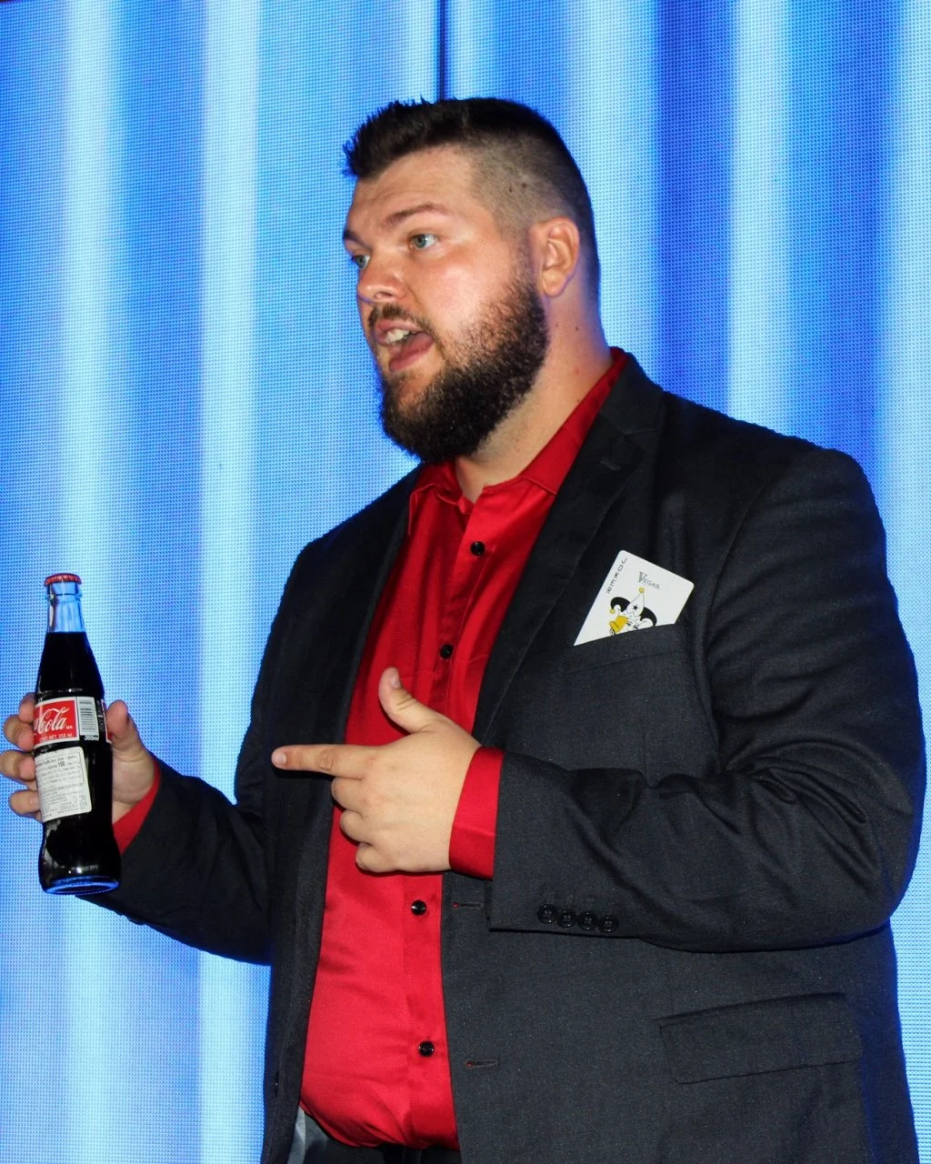 Corporate magician Seth Dreams with a beard and short hair, wearing a dark jacket over a red shirt, is holding a Coca-Cola bottle with his right hand and pointing to the bottle with his left index finers.