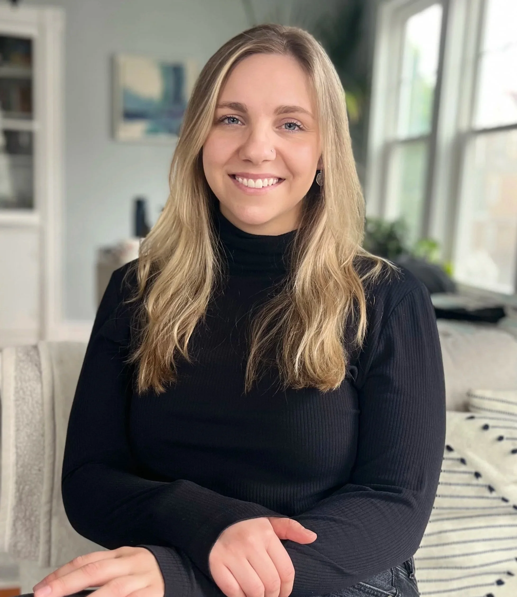 A young woman with long blonde hair smiling, wearing a black turtleneck sweater, sitting in a bright, modern living room with large windows and a painting on the wall.