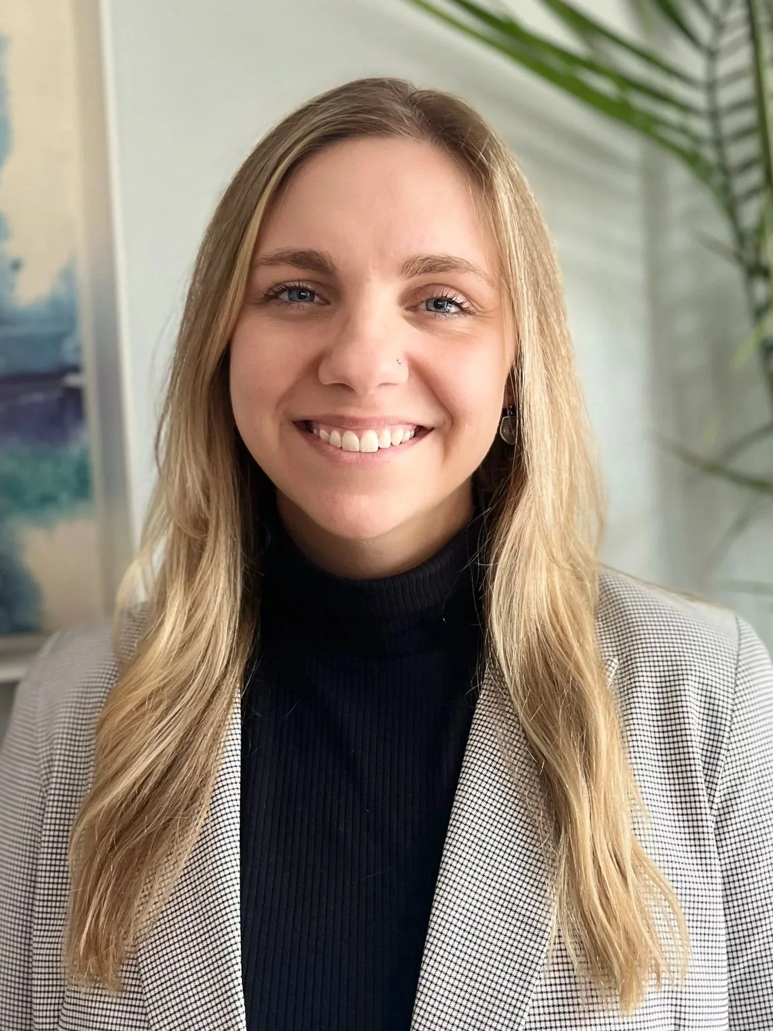 A smiling woman with blonde hair, blue eyes, and a nose piercing, wearing a black turtleneck and a checkered blazer, sitting in a room with artwork and a green plant in the background.