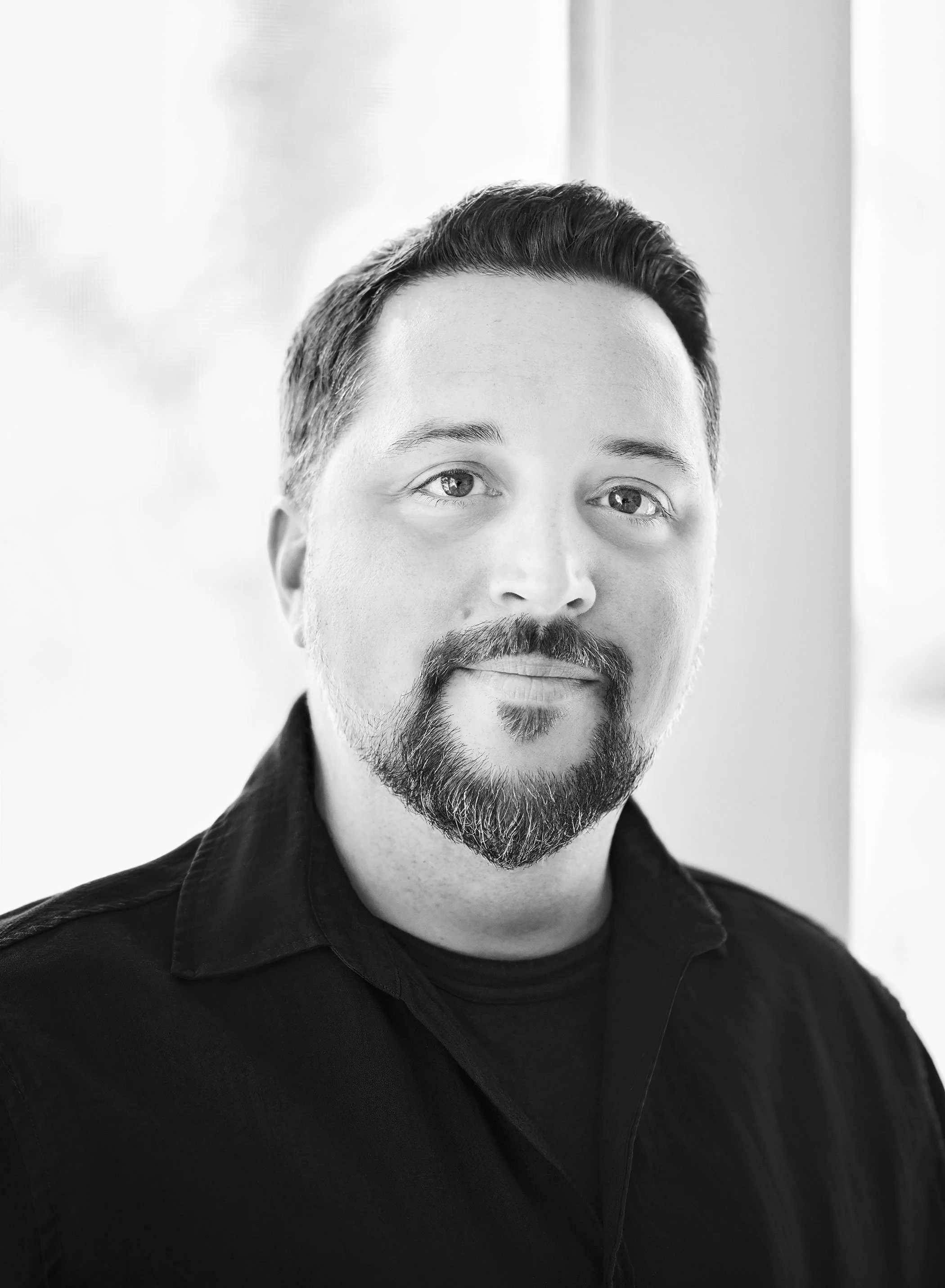Black and white portrait of a man with short dark hair, a beard, and a mustache. He is wearing a dark collared shirt and standing in front of a blurred background with a light source coming from the side.