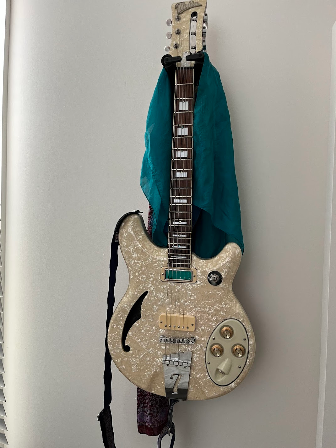 An electric guitar with a pearlescent finish hanging on a wall. The guitar has a semi-hollow body, a distinctive F-hole on the side, gold control knobs, and a black strap. A teal cloth is draped over the neck of the guitar.