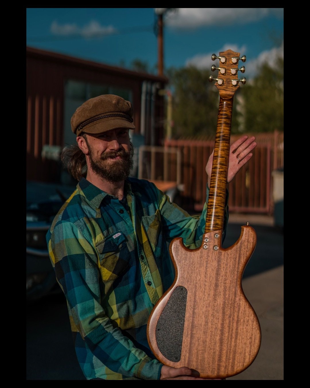 A man with a beard and mustache wearing a brown cap and a plaid shirt holds an electric guitar with a wooden finish outdoors in sunlight.