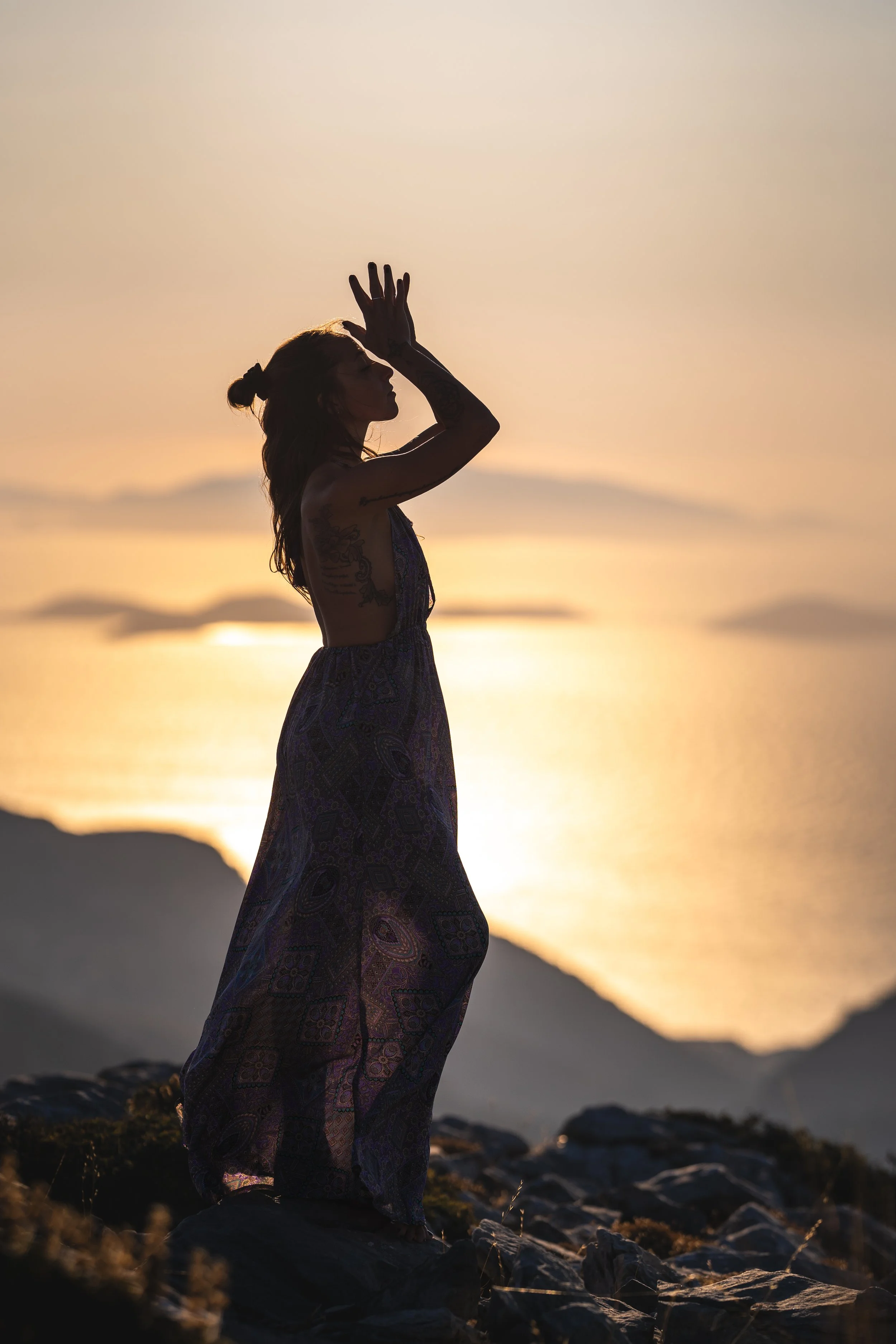 Eine Frau steht auf einem Felsen während eines Sonnenuntergangs am Strand, mit einer Hand an der Stirn, im Schatten lunarischer Silhouette.