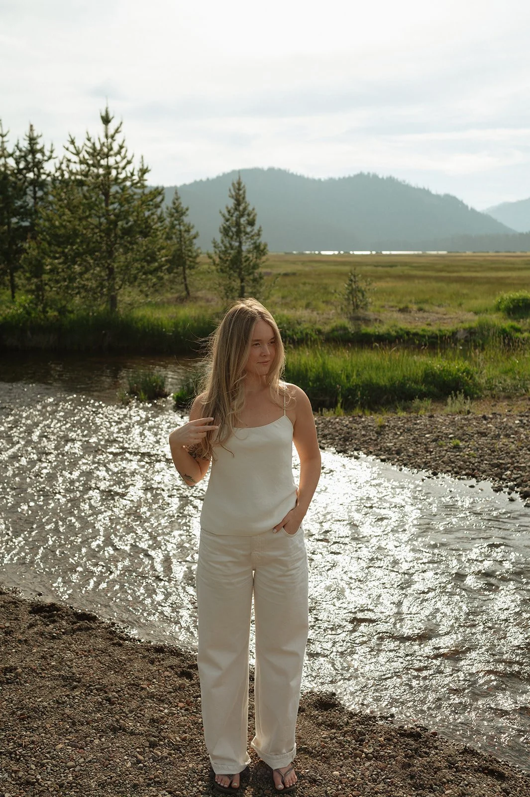 A young woman with long blonde hair wearing a white tank top and white pants stands on a rocky riverbank with water reflecting sunlight, surrounded by green grass, trees, mountains, and a cloudy sky in the background.
