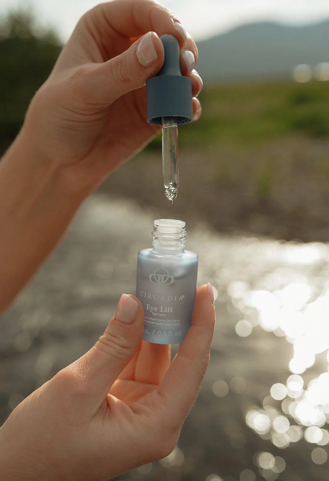 Person holding a bottle of eye lift serum with a dropper, outdoors near a body of water with sunlight reflecting off the surface.
