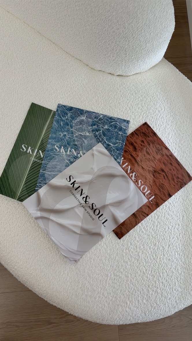 Four brochures for Skin & Soul Corrective Skin Studio on a white textured chair. The brochures have different backgrounds: green leaf, blue water, white marble, and brown sand.
