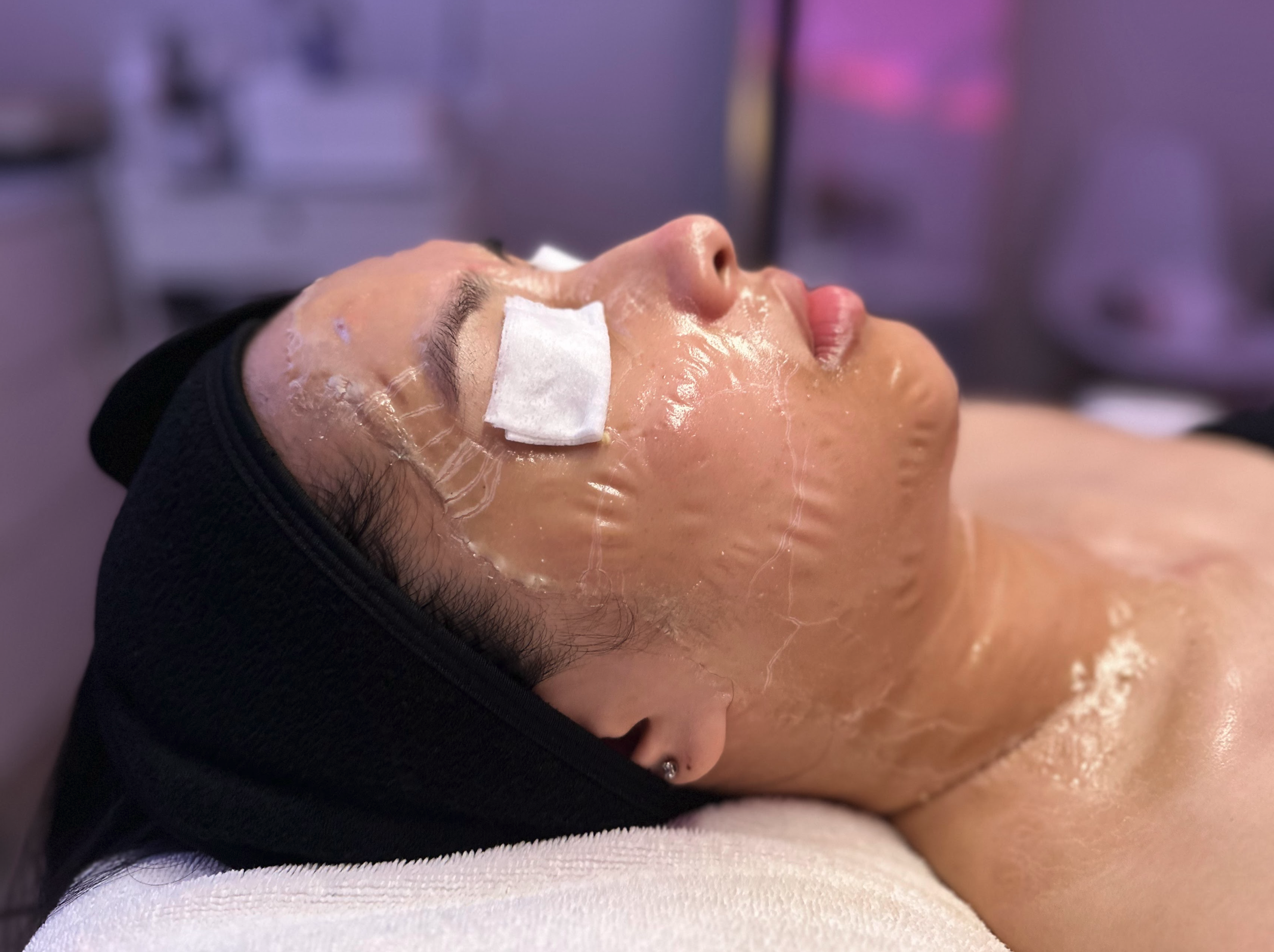 Close-up of woman receiving a facial treatment with cleansing mask, lying down with eye patches, in a spa or clinic setting.