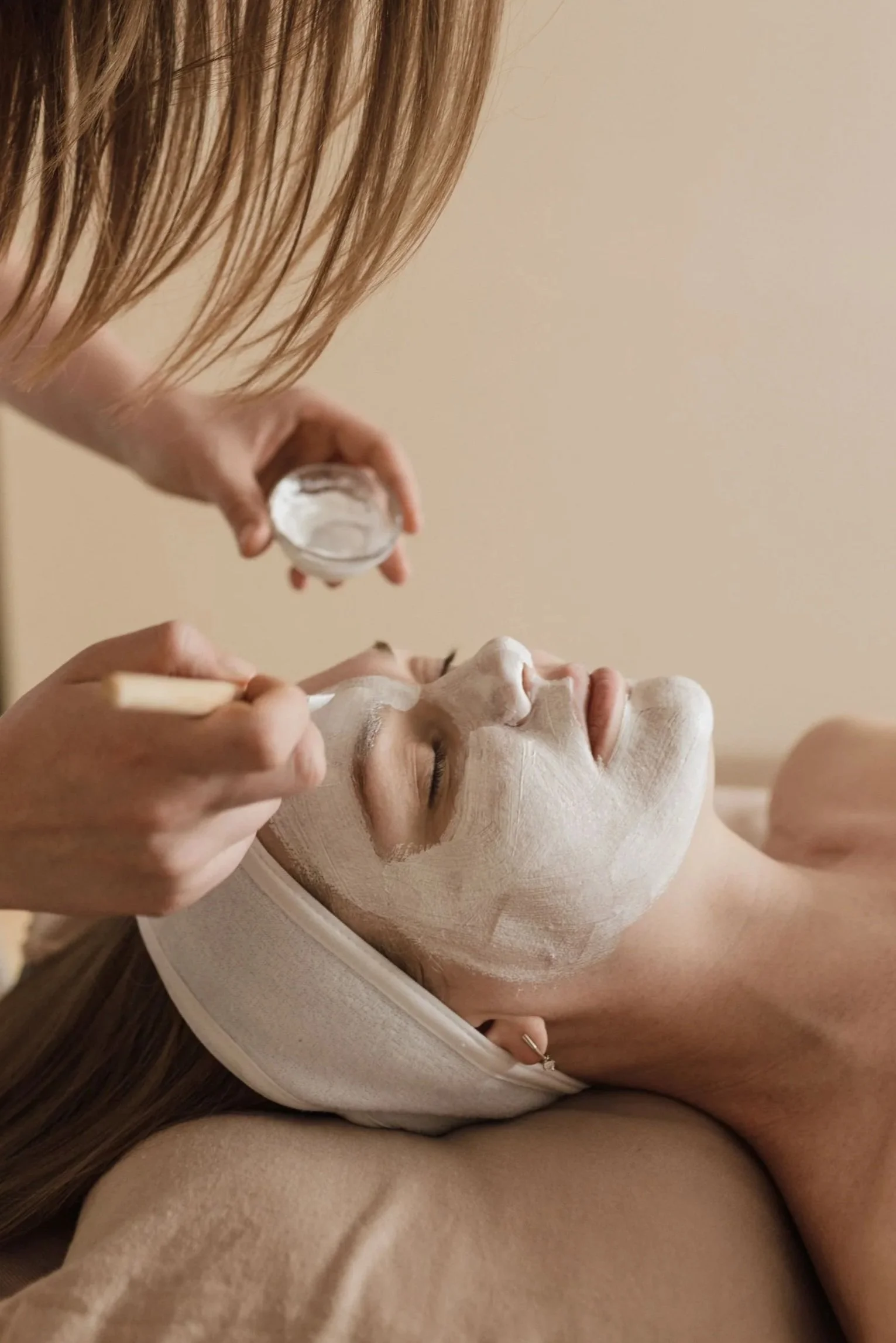 Person lying down with closed eyes while someone applies a facial mask using a small jar, with part of the person's face painted with the mask and a towel around the neck.