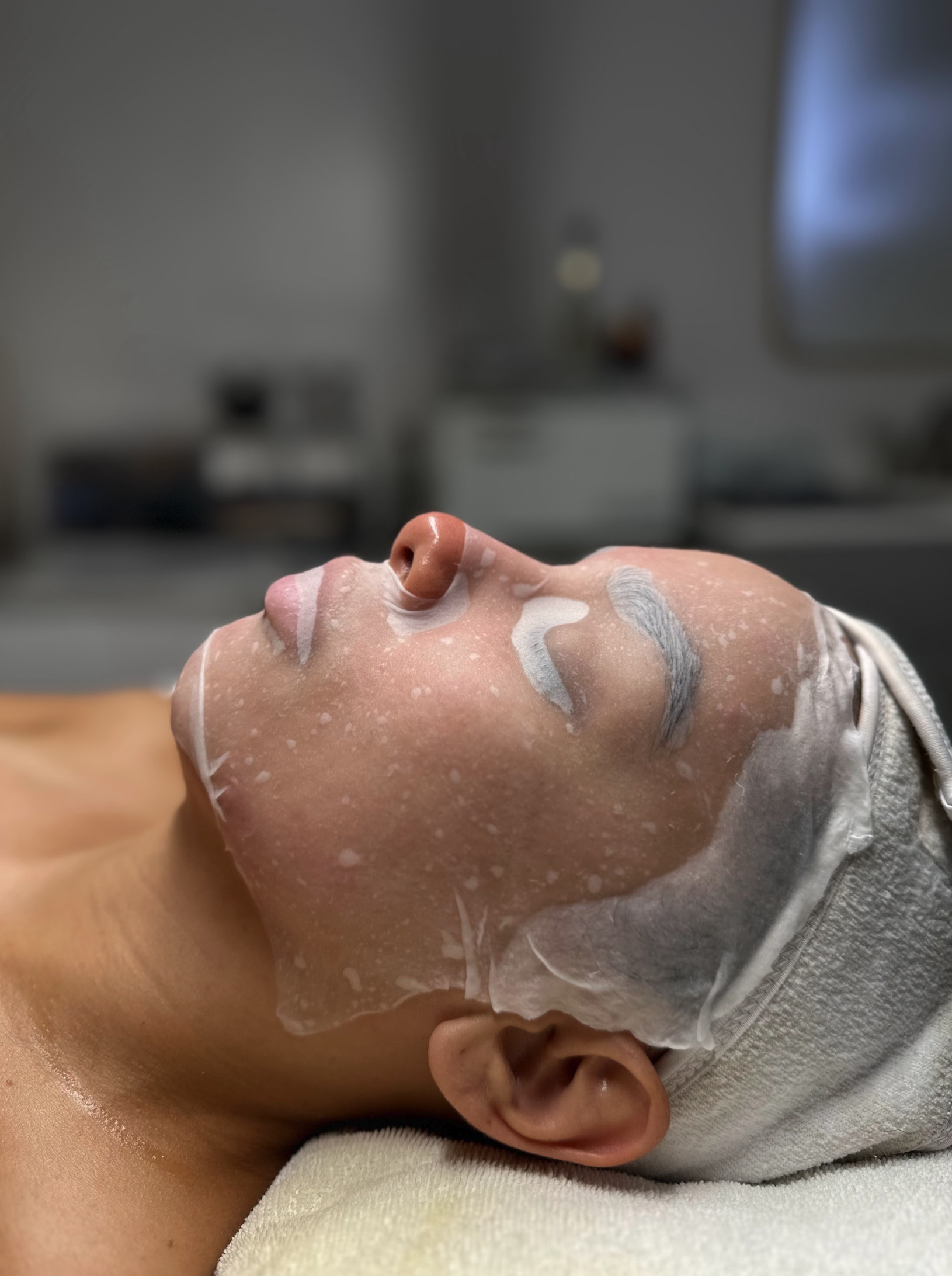 Close-up of a person's face with a facial mask applied, lying down with eyes closed, in a skincare or spa setting.