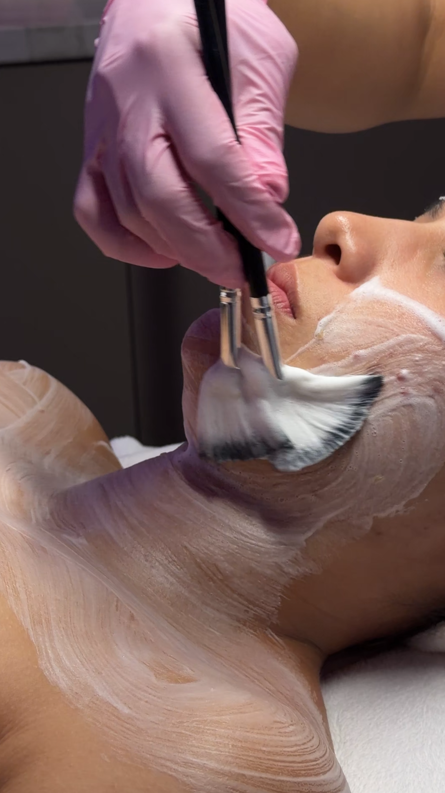 A person lying on a spa or massage table receiving a facial treatment with a brush, with a skincare mask applied to their face.