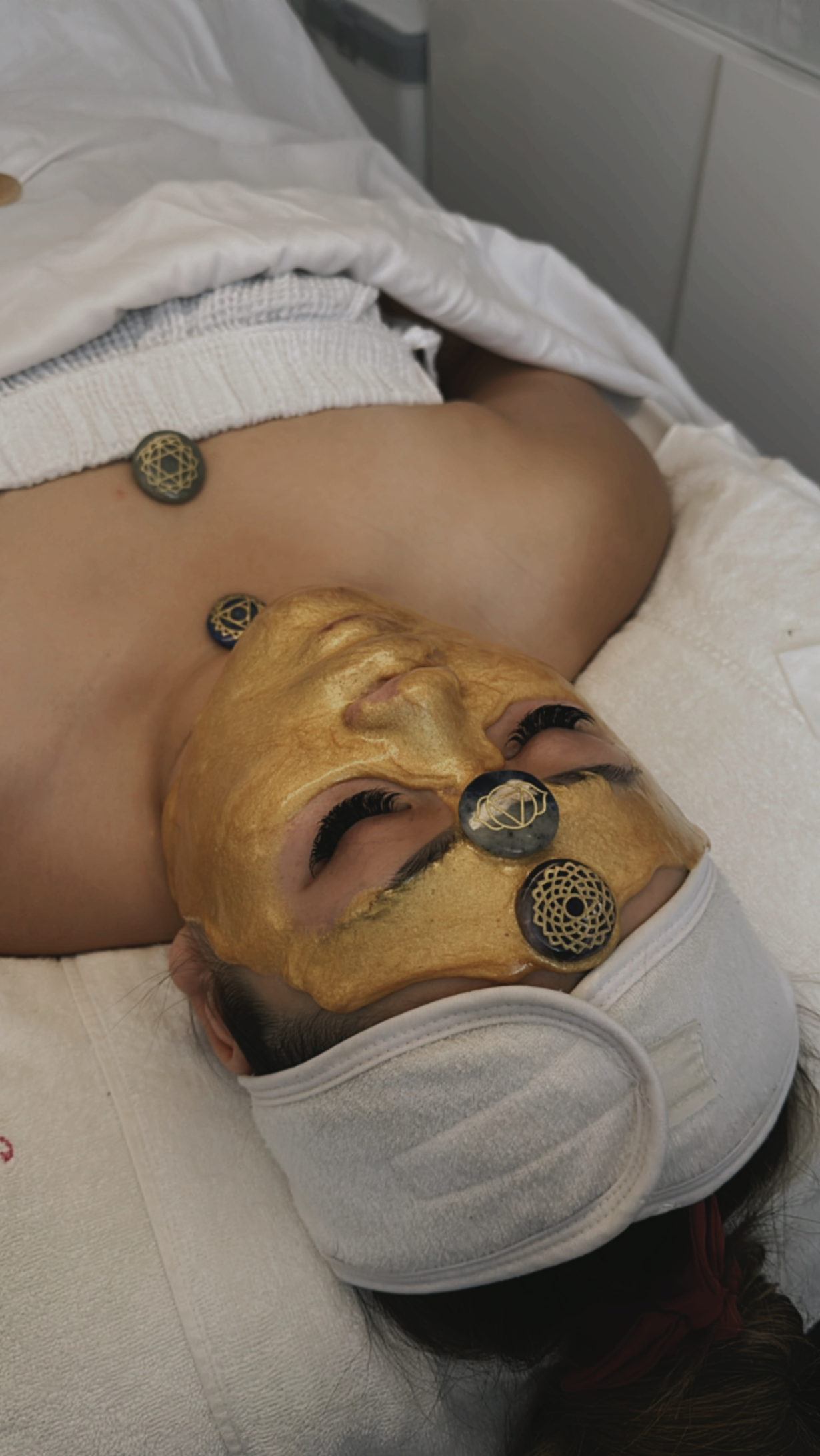 A woman lying on a bed with a gold facial mask applied to her face. She has black eyelashes and eyes open. She wears a white towel or headband, with decorative black and gold facial disks on her forehead and chin.