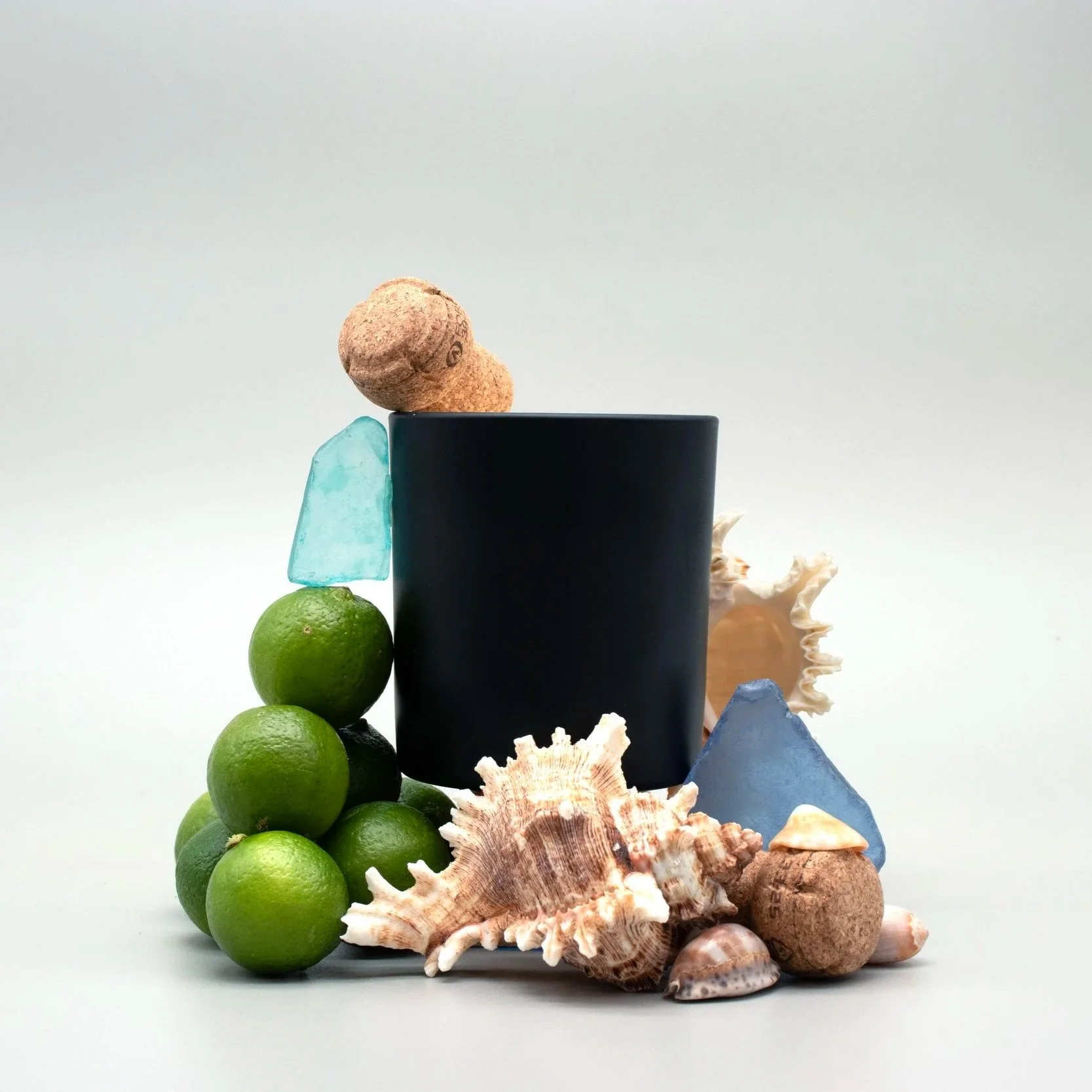 Sculptural still life with a clean burning candle, key limes, champagne corks, sea glass and shells.