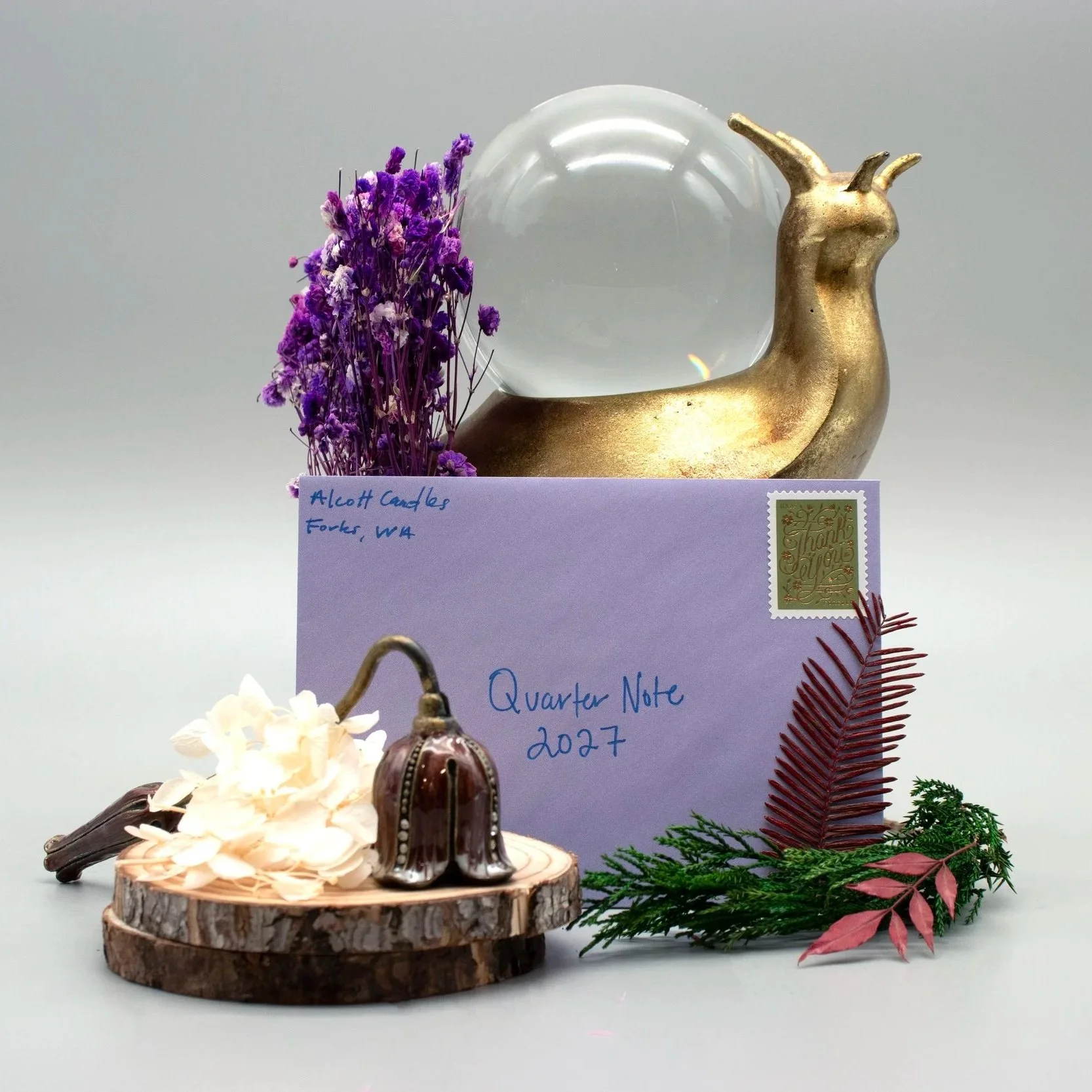 Still life with a letter that is addressed to Quarter Note 2027, greenery, purple flowers, an antique candle snuffer and pine branches.