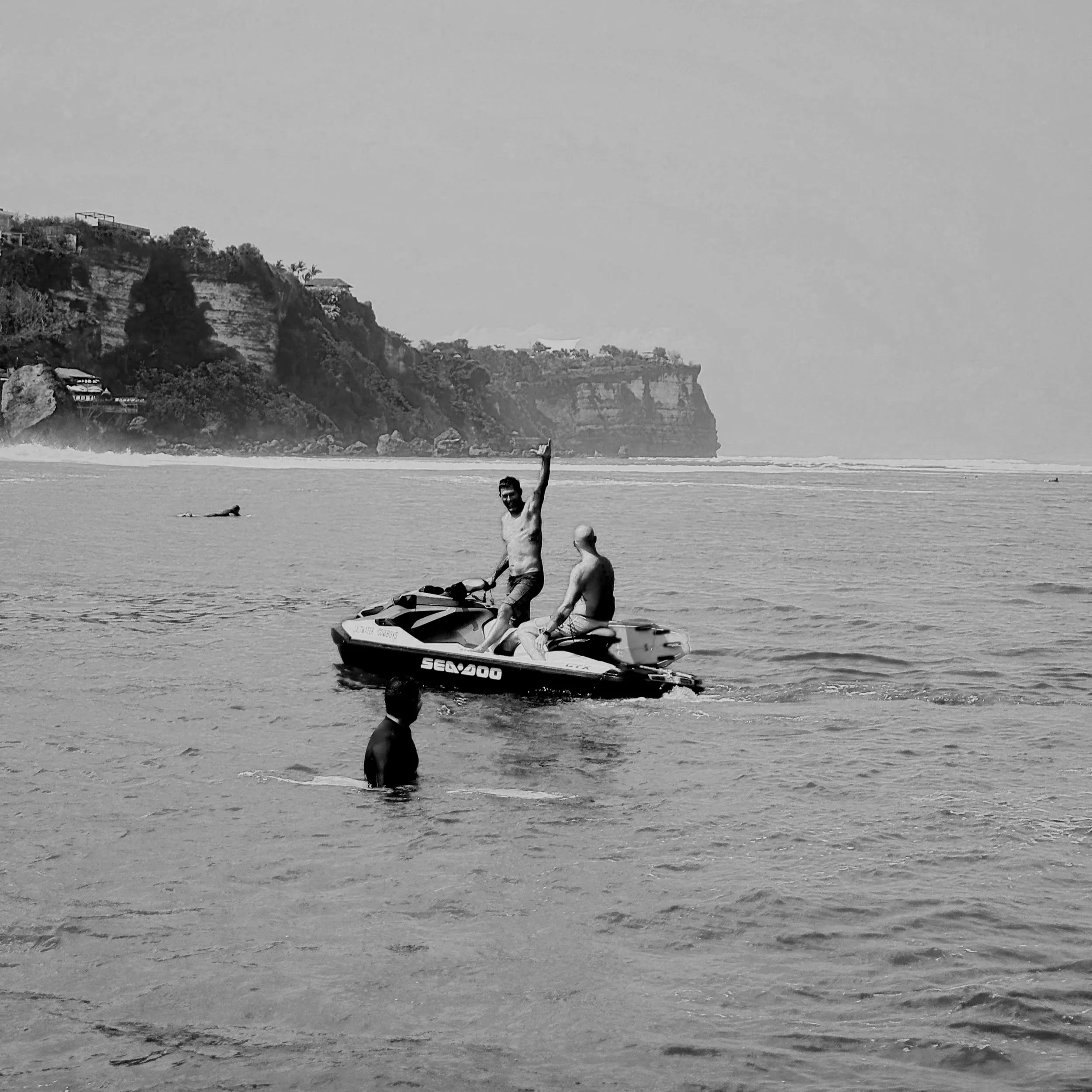 Two men on a yellow Sea-Doo jet ski in the ocean with a man swimming nearby. A scenic coastline with cliffs and green trees in the background under a blue sky.