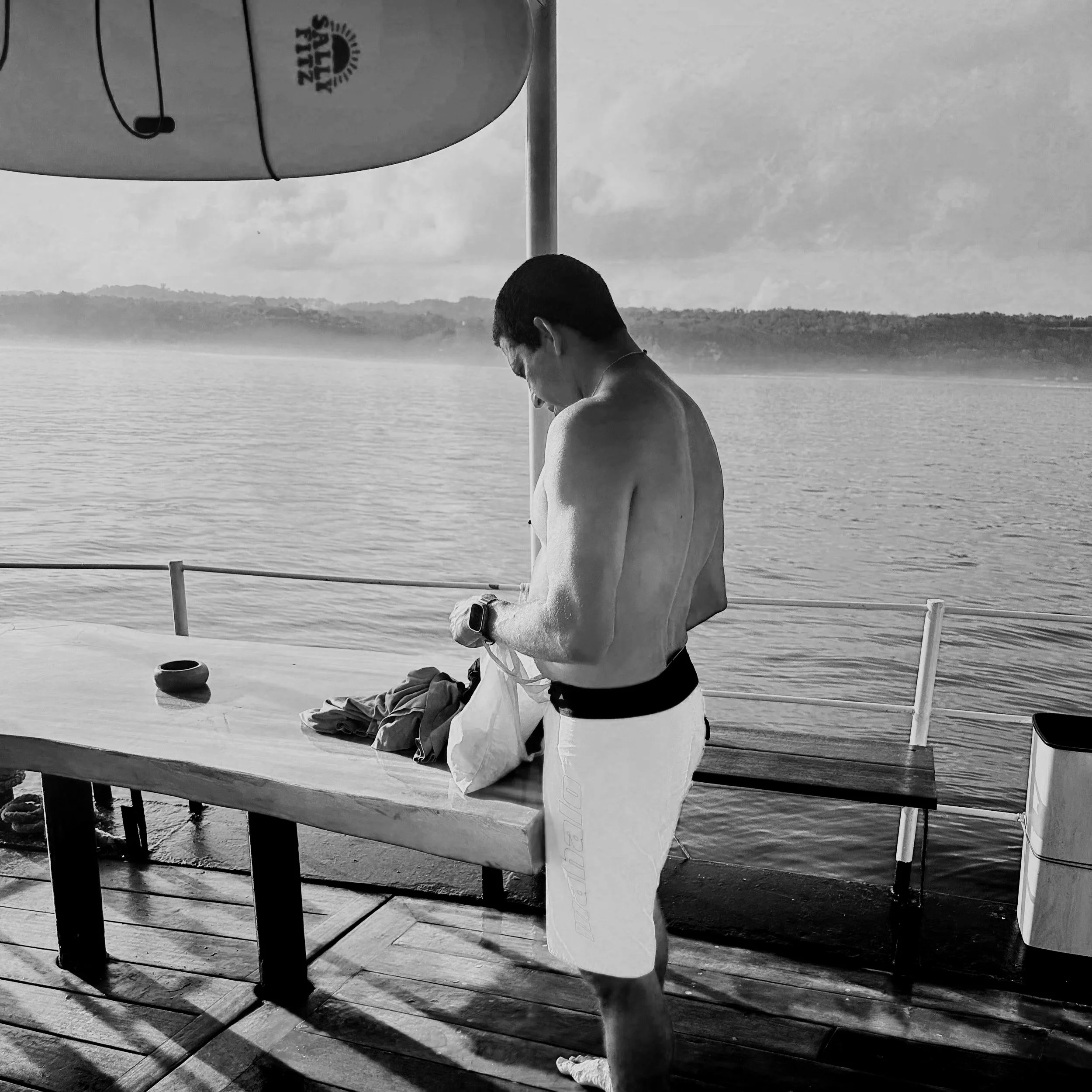 A man in orange swim trunks stands on the deck of a boat near the water, looking down and holding a towel or cloth. The boat has a wooden deck and a large red life preserver hanging from a pole. The water and distant shoreline are visible in the back