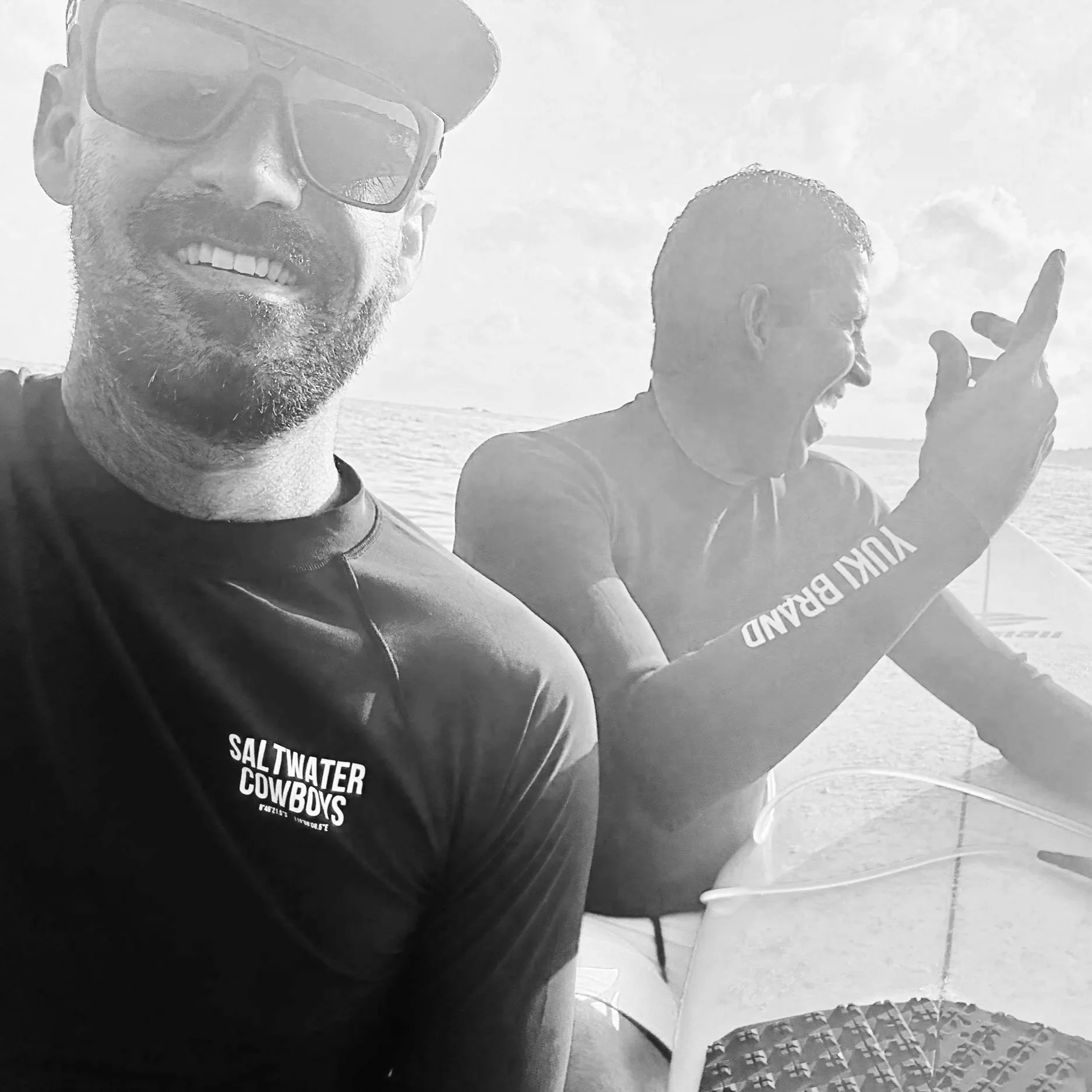 Two men wearing black Saltwater Cowboys shirts, one smiling at the camera and the other laughing, on a boat in the water with a bright sky and clouds in the background.