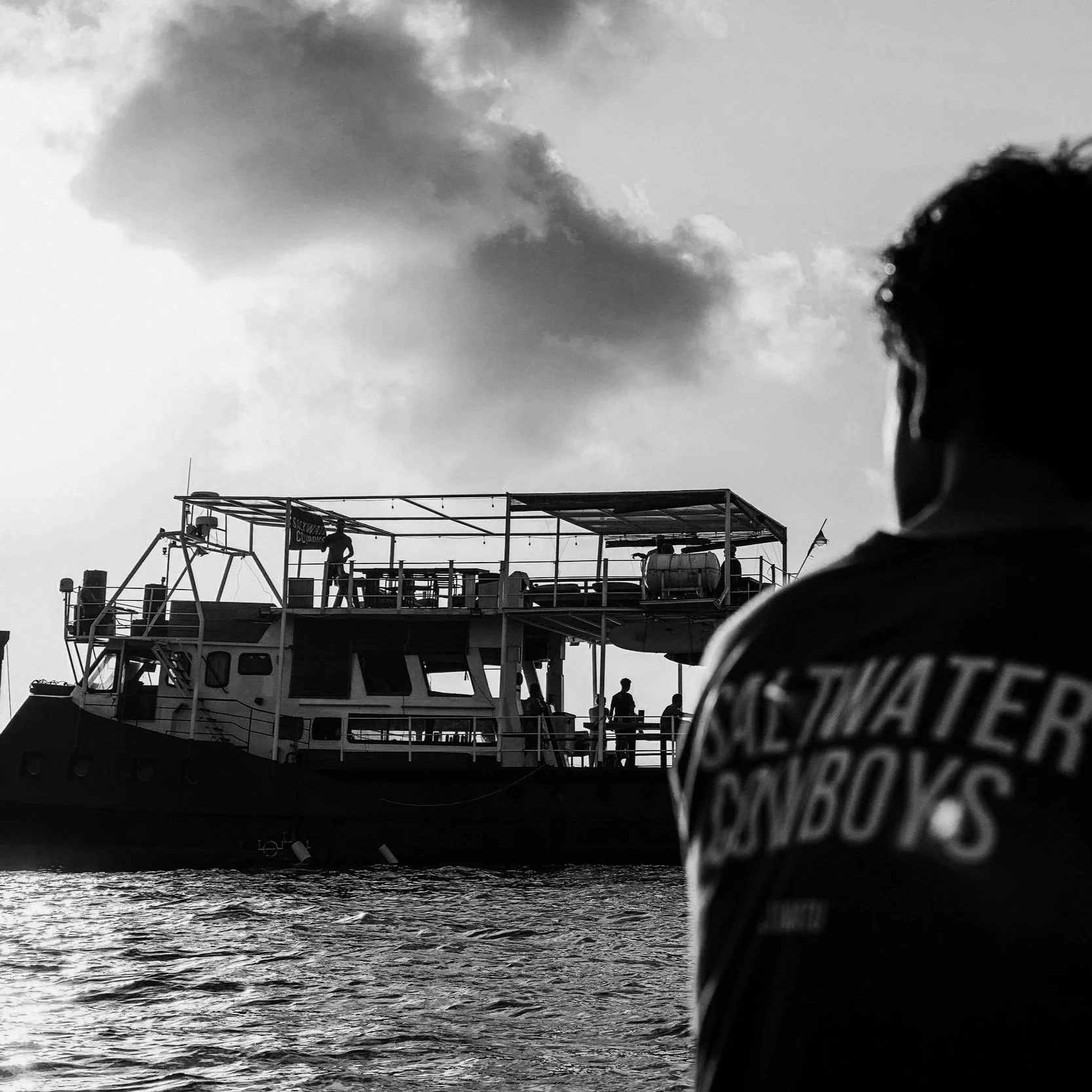 A person with 'Saltwater Cowboys' on the back of their shirt watches a boat on the water during sunset.